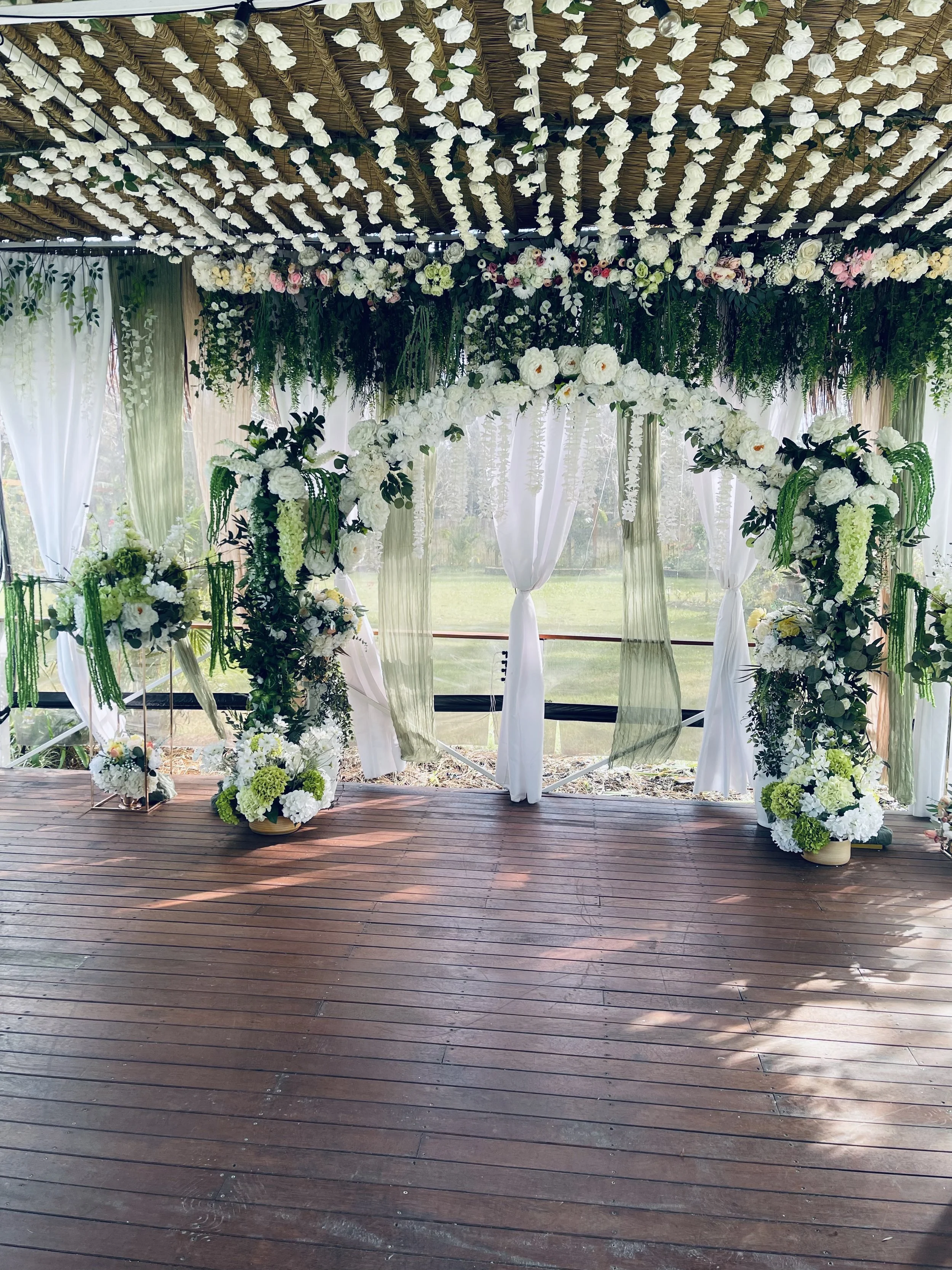 A wedding ceremony setup with an arch decorated with white flowers and greenery, surrounded by hanging floral arrangements from a thatched roof ceiling, with sheer curtains and potted floral arrangements on a wooden platform.