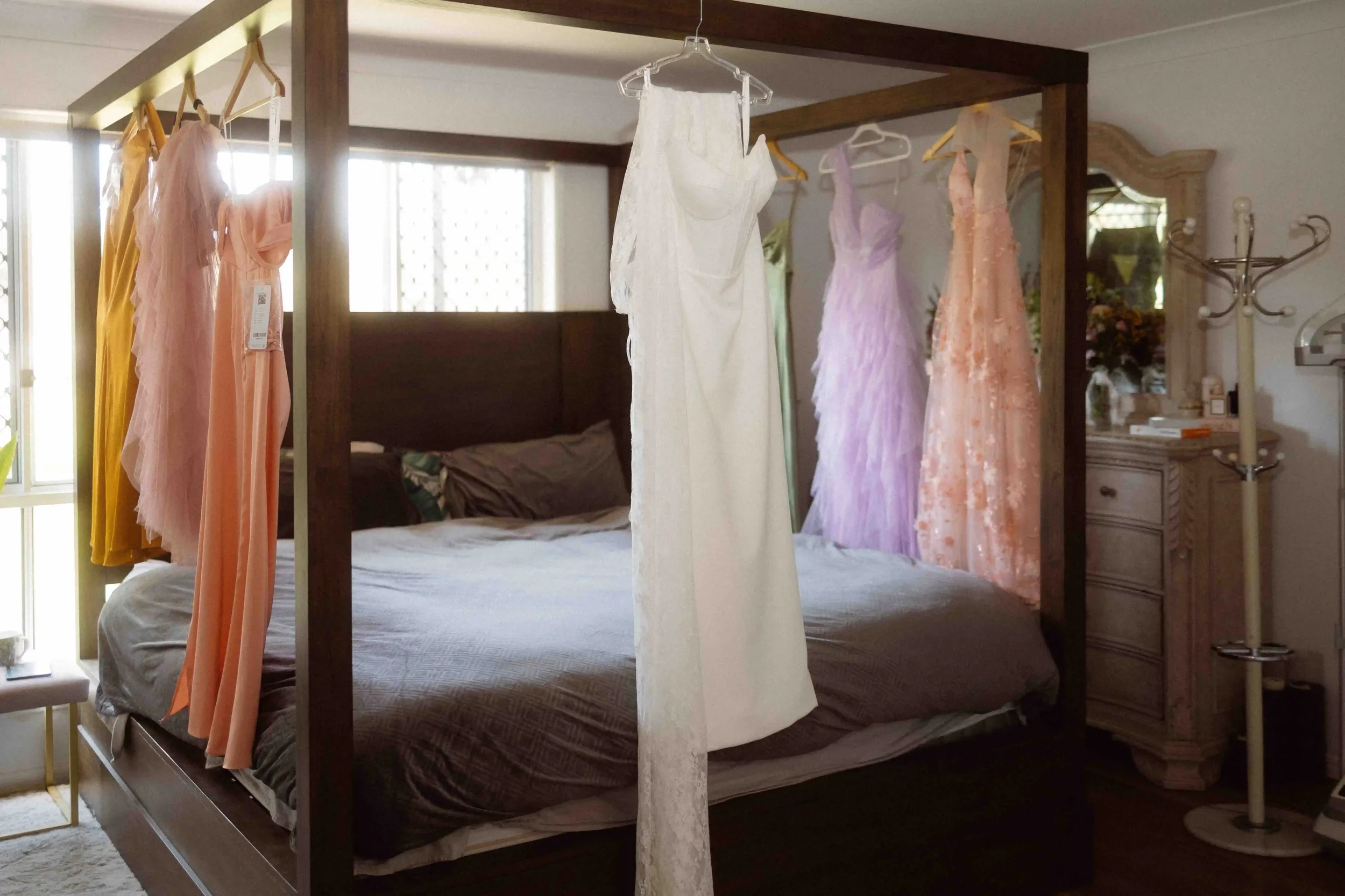 A bedroom with a four-poster bed and a sheer curtain hanging from the frame, with various pastel-colored dresses hanging on the bed's frame, and a nightstand with a mirror and lamp nearby.