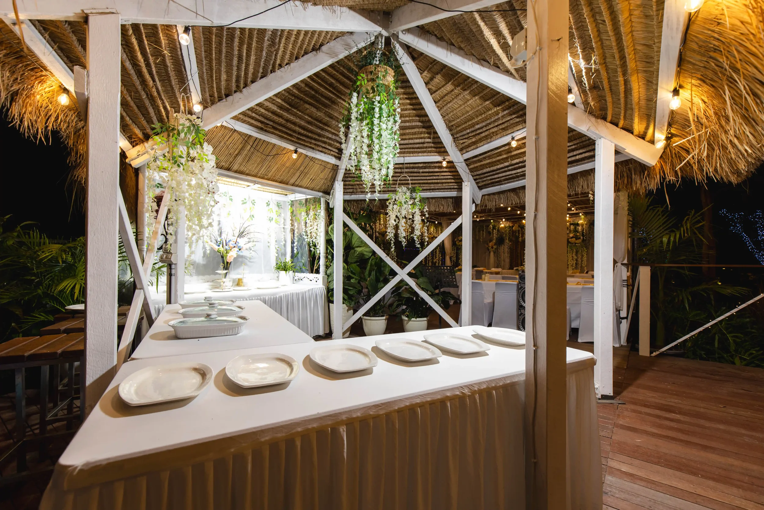 A decorated outdoor dining area with a thatched roof, hanging plants, string lights, and tables set with white plates.