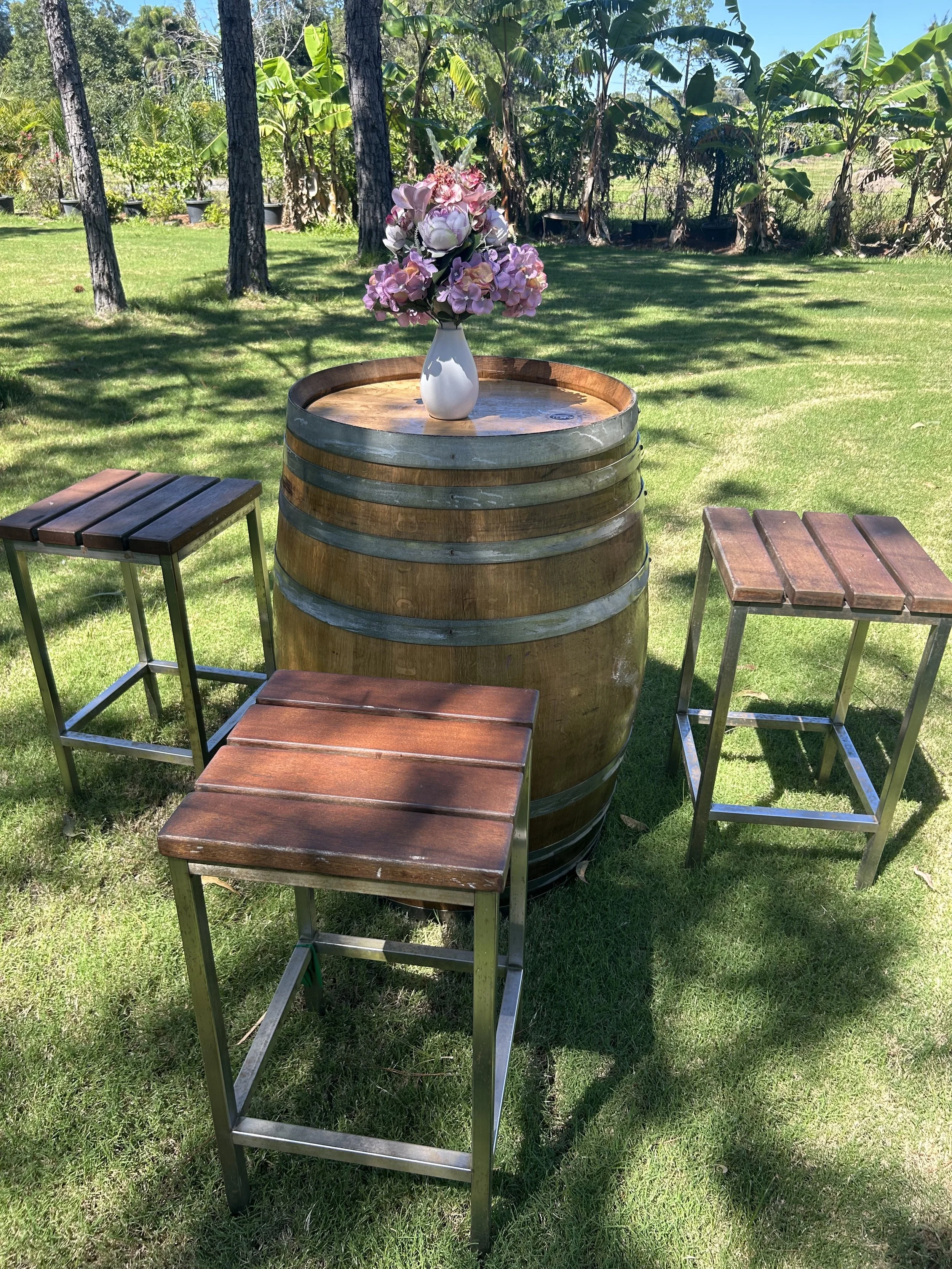 A wooden barrel with a white vase holding pink and purple flowers on top, set outdoors with four wooden stools around it, on a grassy area with trees and plants in the background.