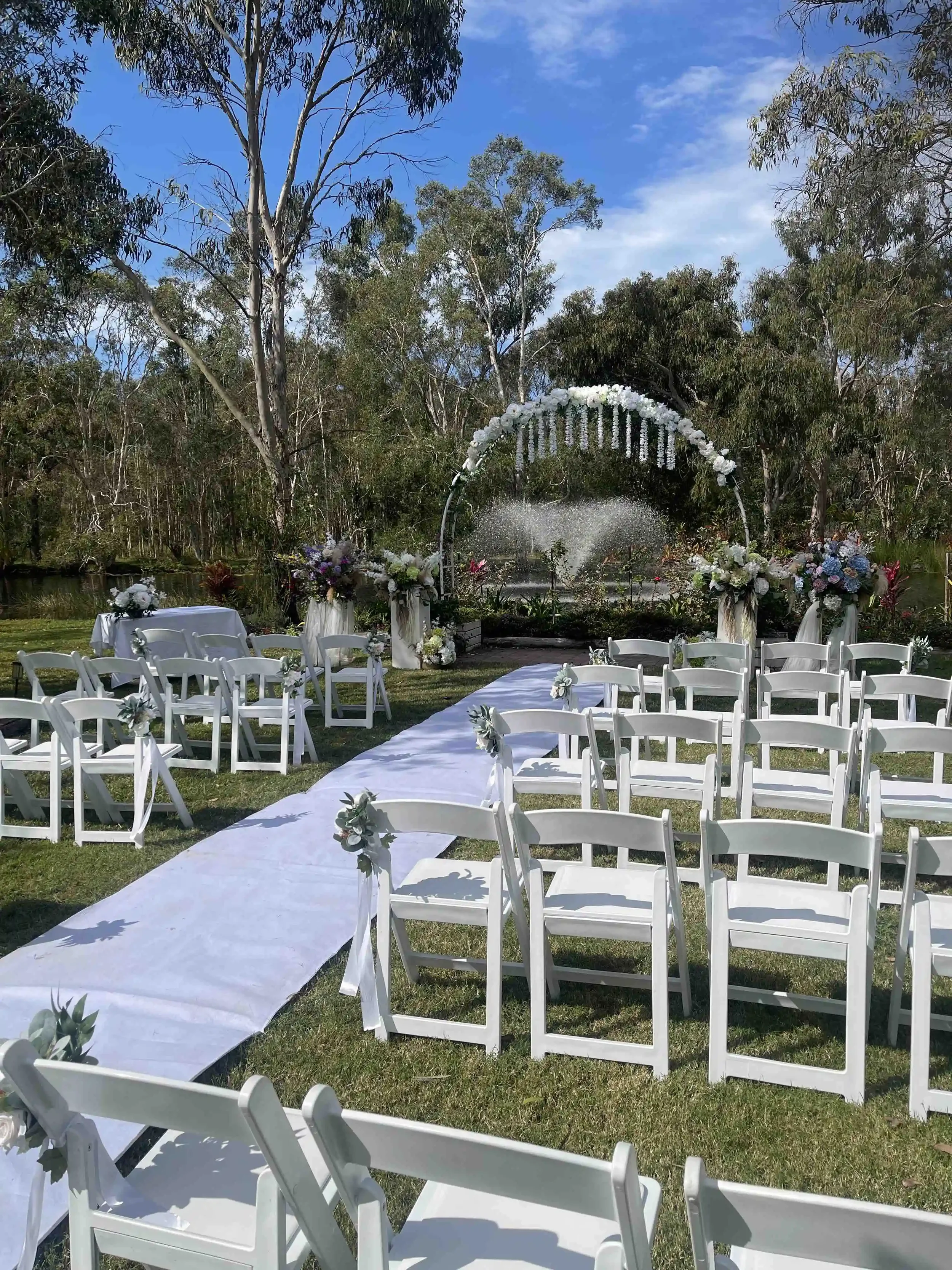 Outdoor wedding setup with white chairs, decorated aisle, floral arch, and floral arrangements, set against a background of trees and a blue sky.