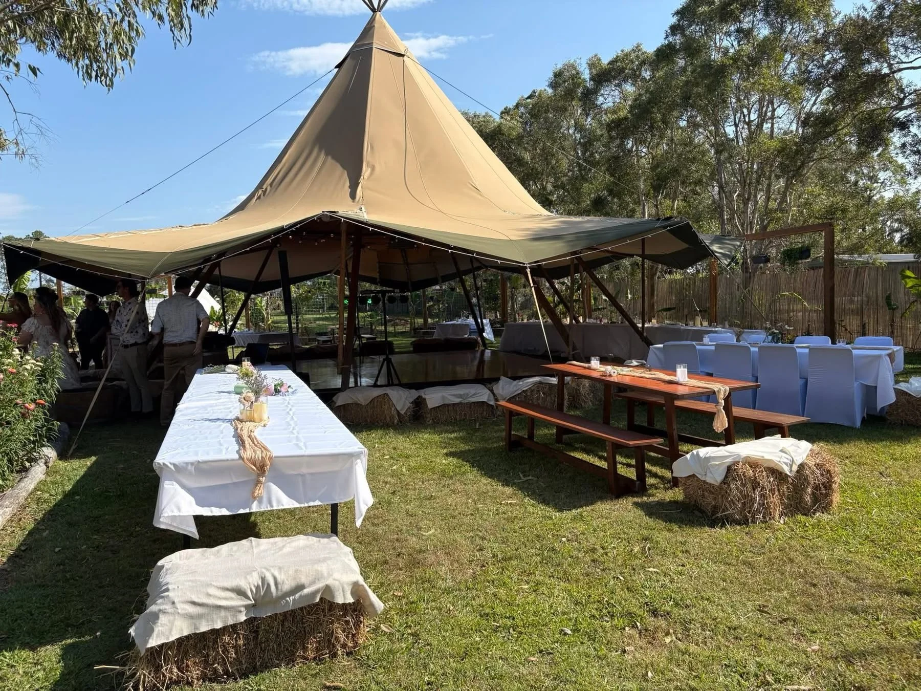 Outdoor wedding reception setup with a large teepee-style tent, long dining tables with white tablecloths, hay bales covered with cloth used as seating, and surrounded by trees.