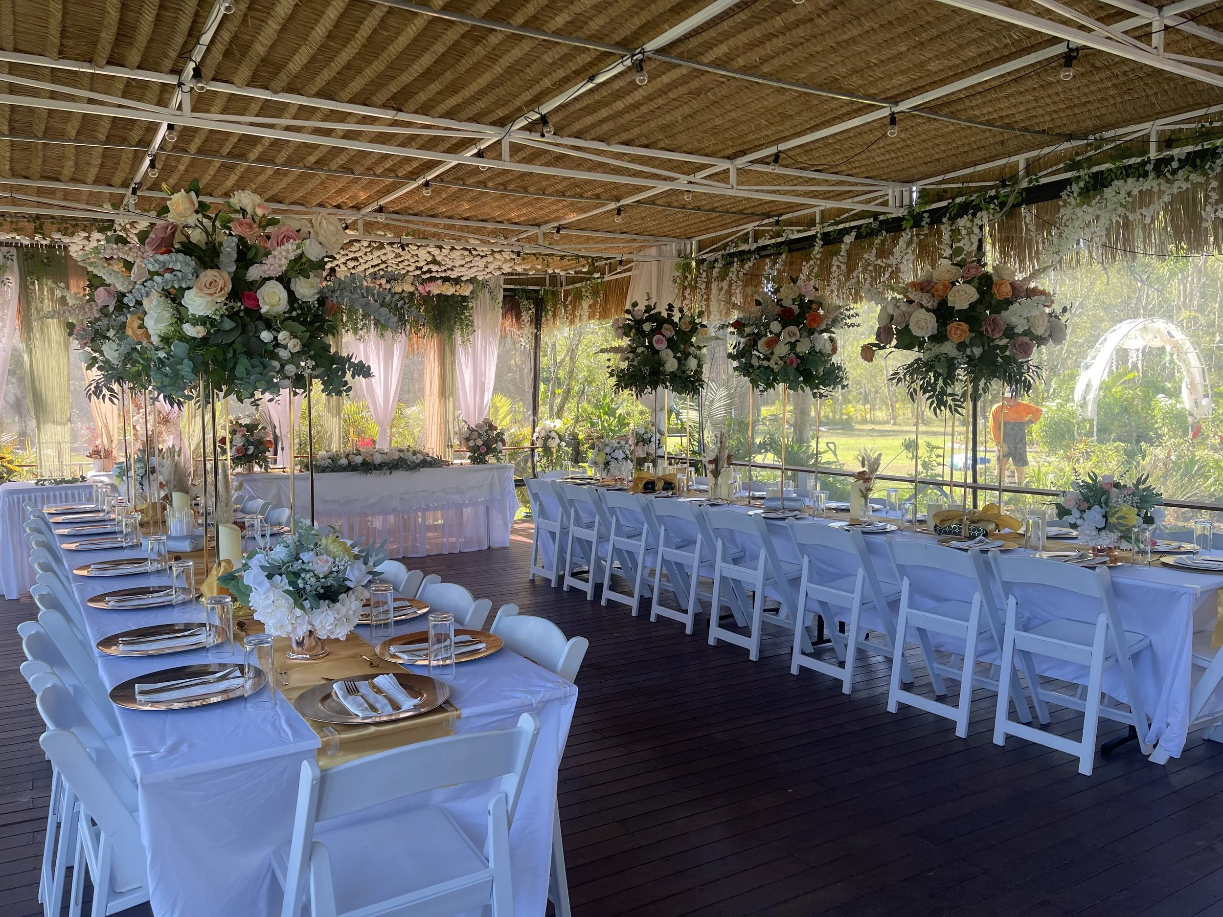 A decorated outdoor event space with white tables and chairs, floral centerpieces, and a wooden ceiling, set up for a celebration or wedding reception.