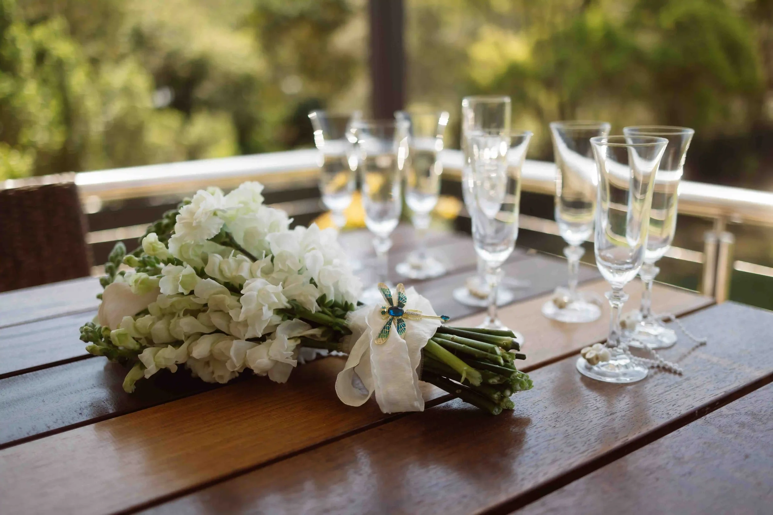 A bouquet of white flowers with a dragonfly pin lies on a wooden table, with several empty glass glasses in the background outdoors.