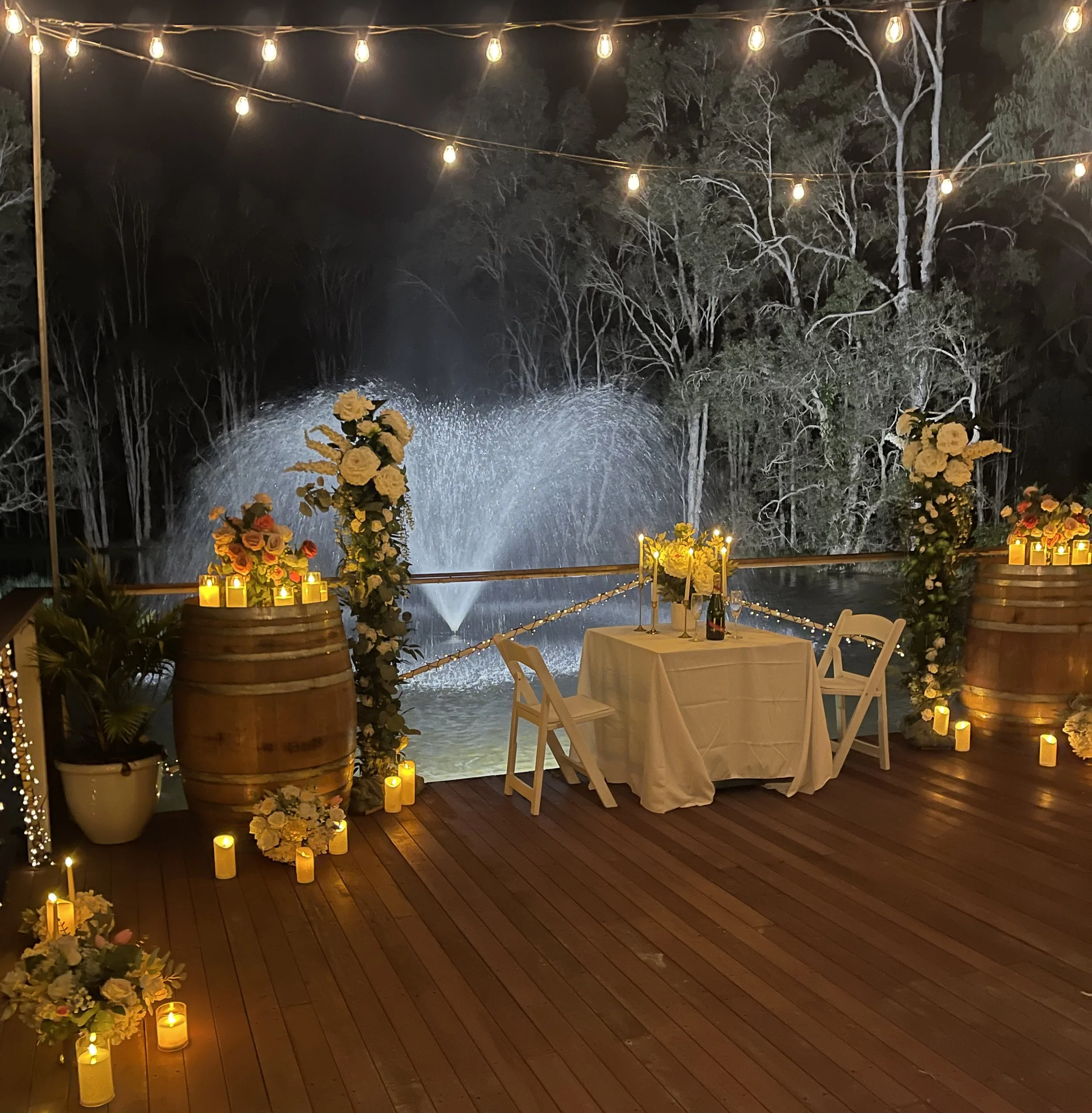 Nighttime outdoor setting with string lights hanging above, surrounded by trees. In the foreground, there are tables with white tablecloths, yellow candles, and floral arrangements, creating a warm, inviting atmosphere.