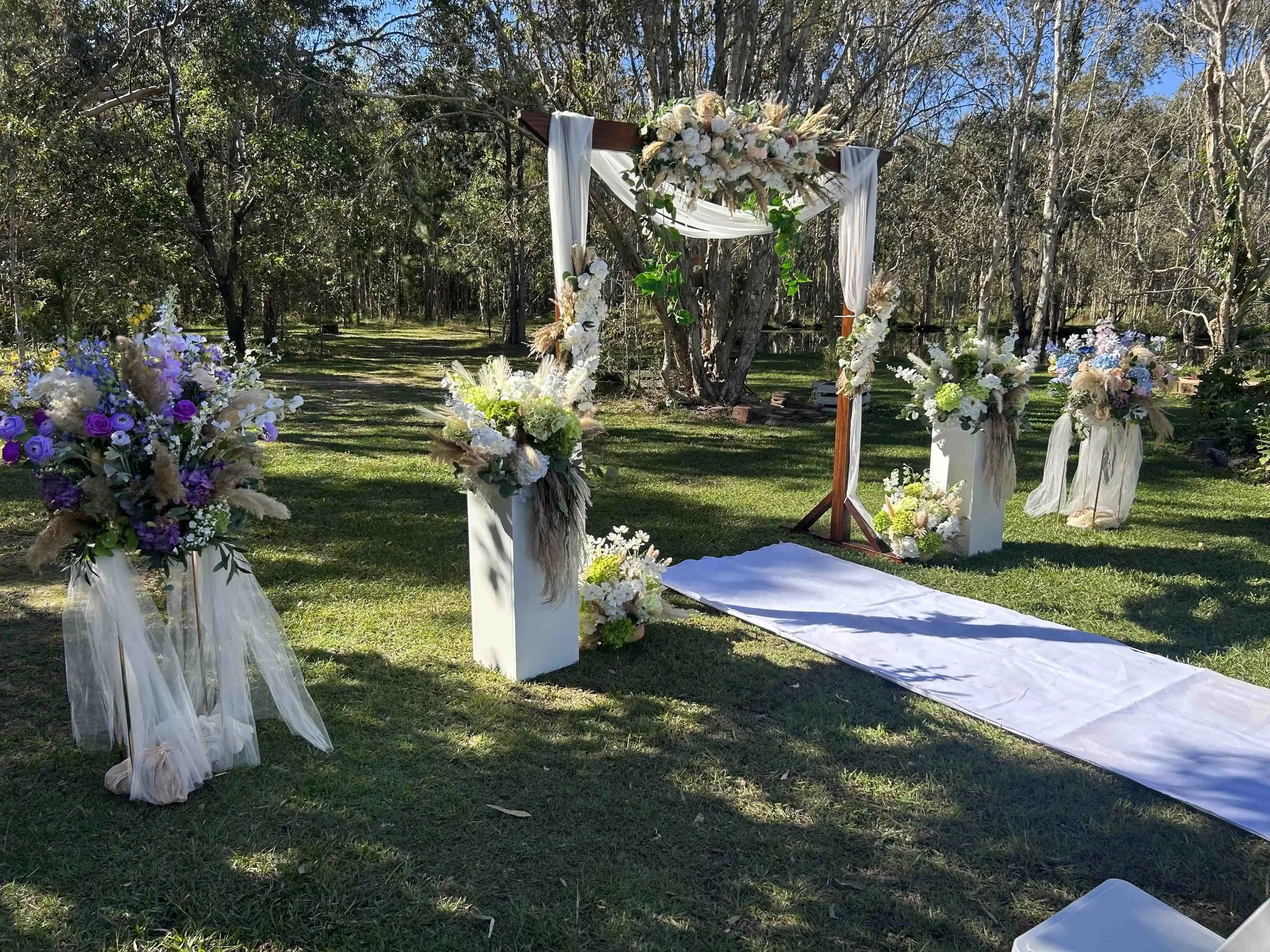 Outdoor wedding arch decorated with flowers and white fabric set up in a grassy area surrounded by trees.