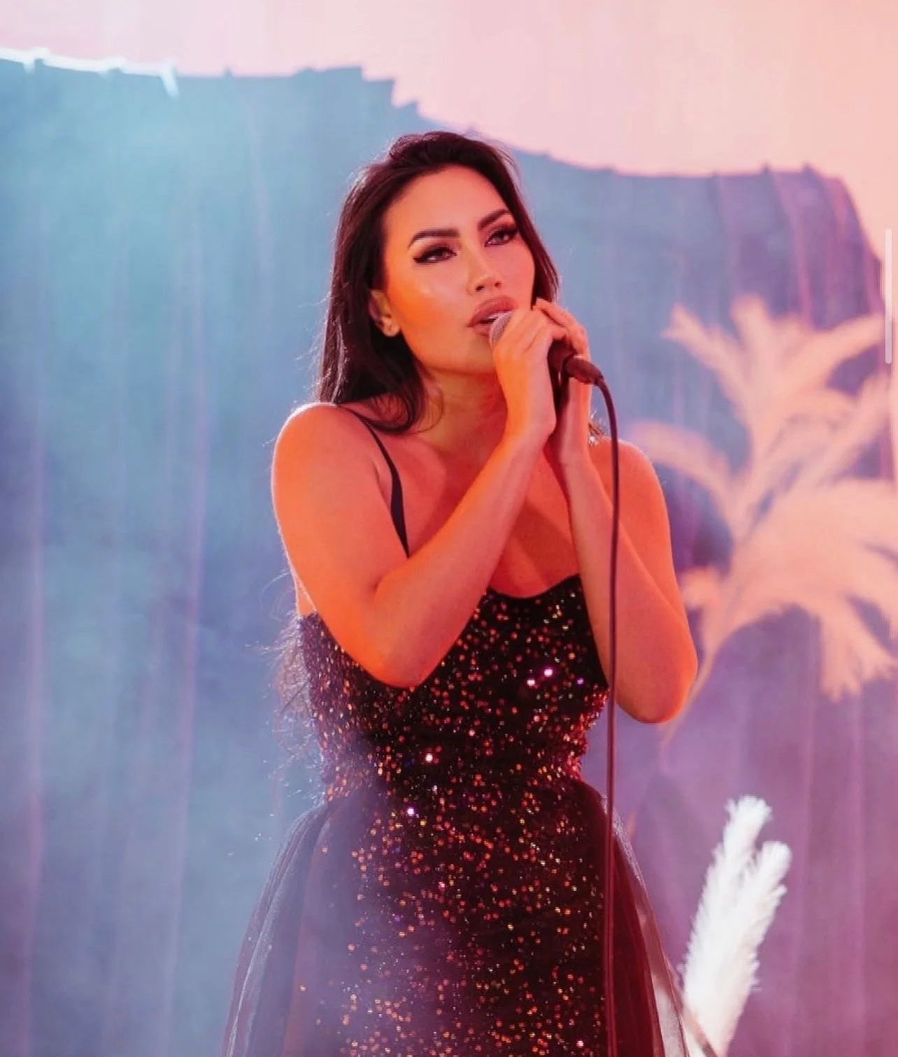 A woman with dark hair and makeup, wearing a black, sparkly, spaghetti-strap dress, singing into a microphone on stage with pink and blue lighting.