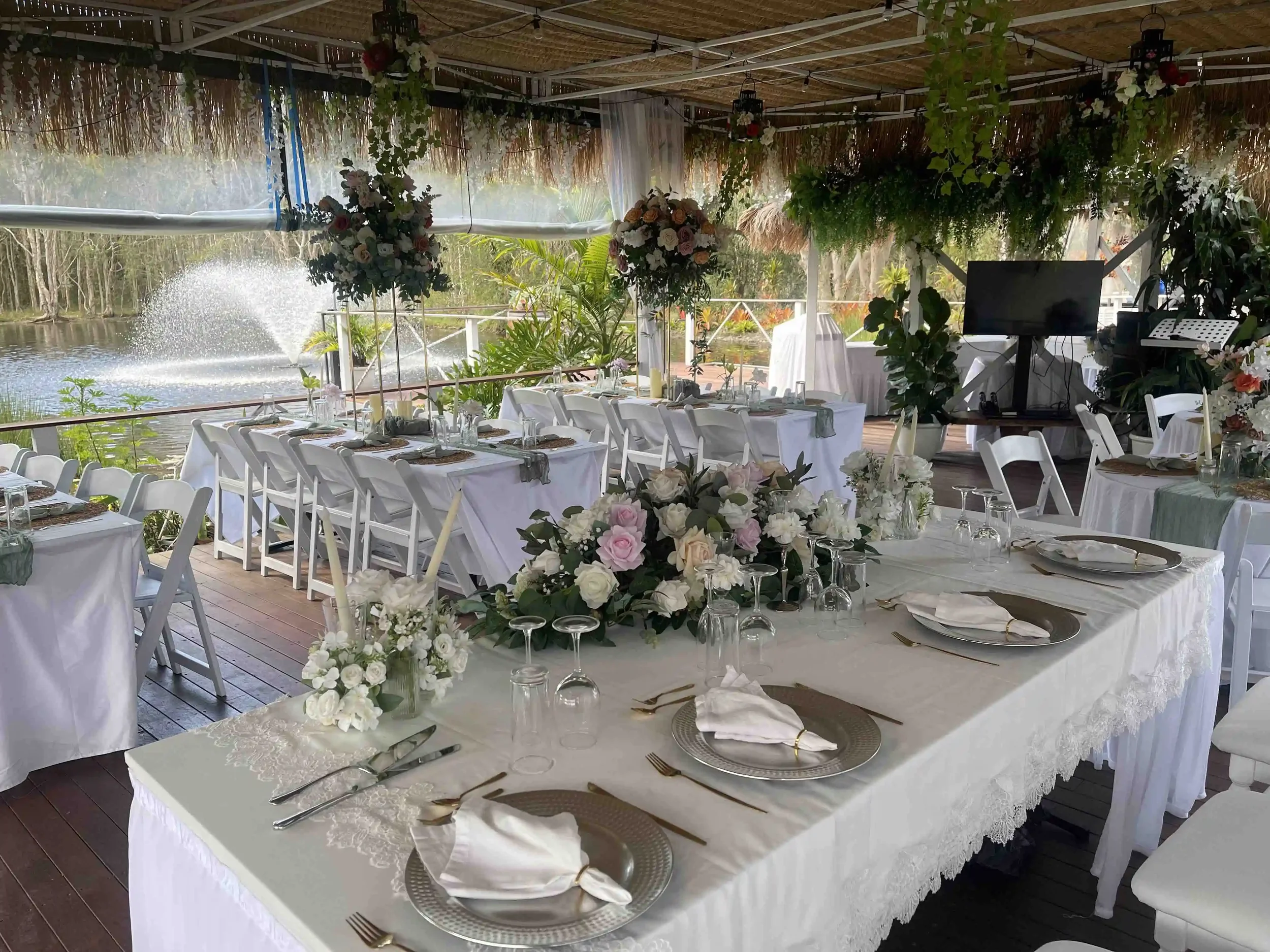 Decorated outdoor dining area with white tablecloths, floral centerpieces, and a view of water with a fountain, surrounded by greenery and trees.