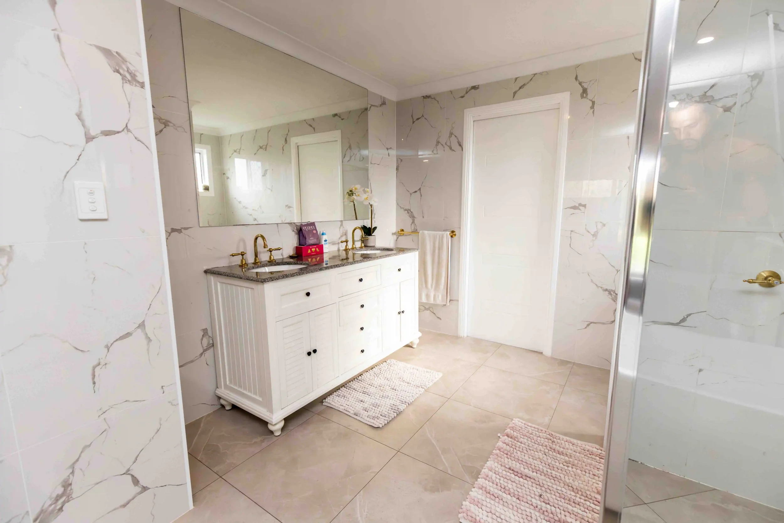 Bathroom with marble walls, a double vanity with gold fixtures, large mirror, white cabinet, and shower with glass door.