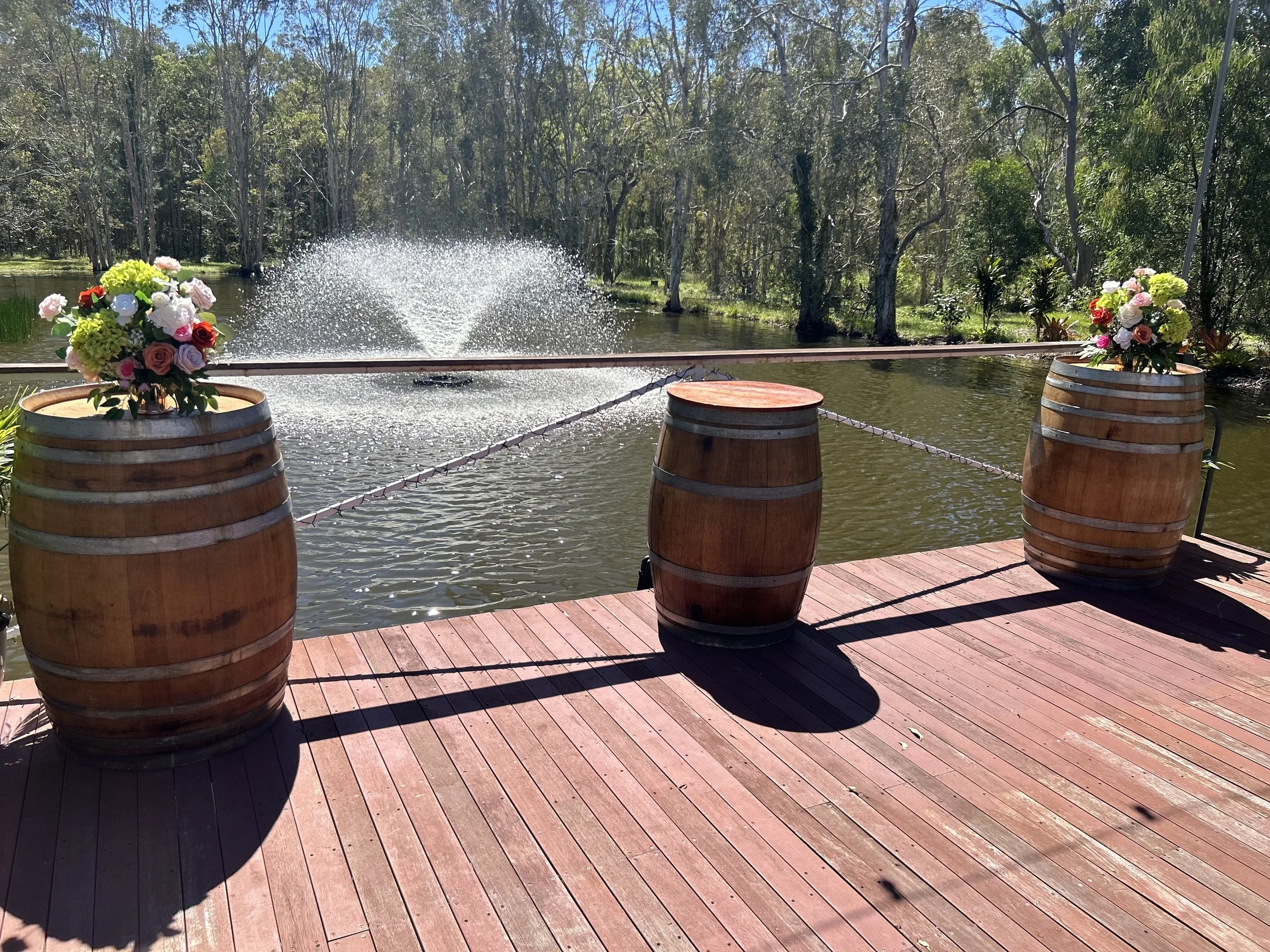 A lakeside scene with three wooden barrels used as planters holding colorful flower arrangements. There is a railing in front of the barrels, a wooden deck, and a lake with trees in the background. The water is calm with a fountain visible.