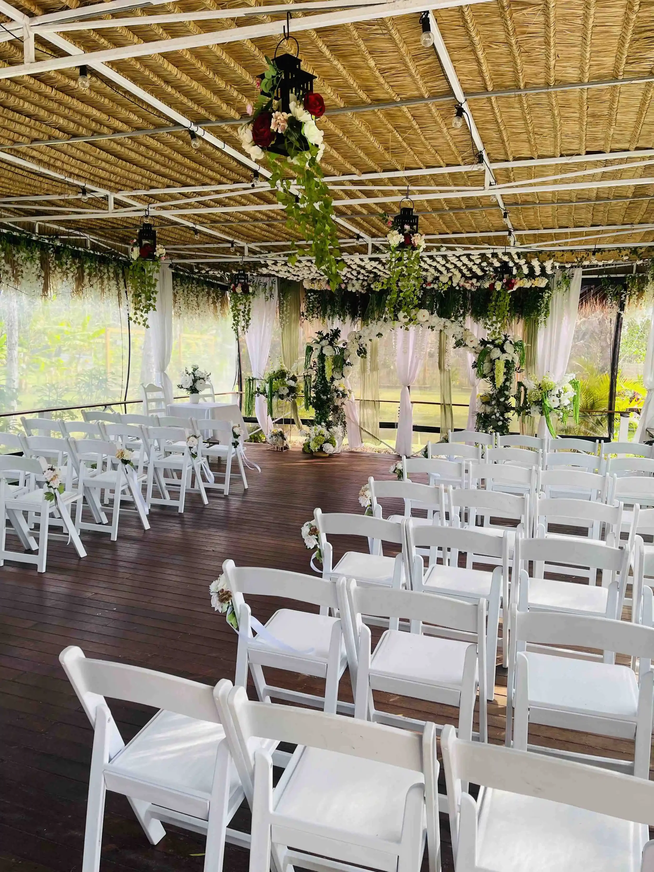 Outdoor wedding setup with white chairs arranged in rows facing an arch decorated with flowers, under a thatched roof ceiling with hanging lanterns and flowers.
