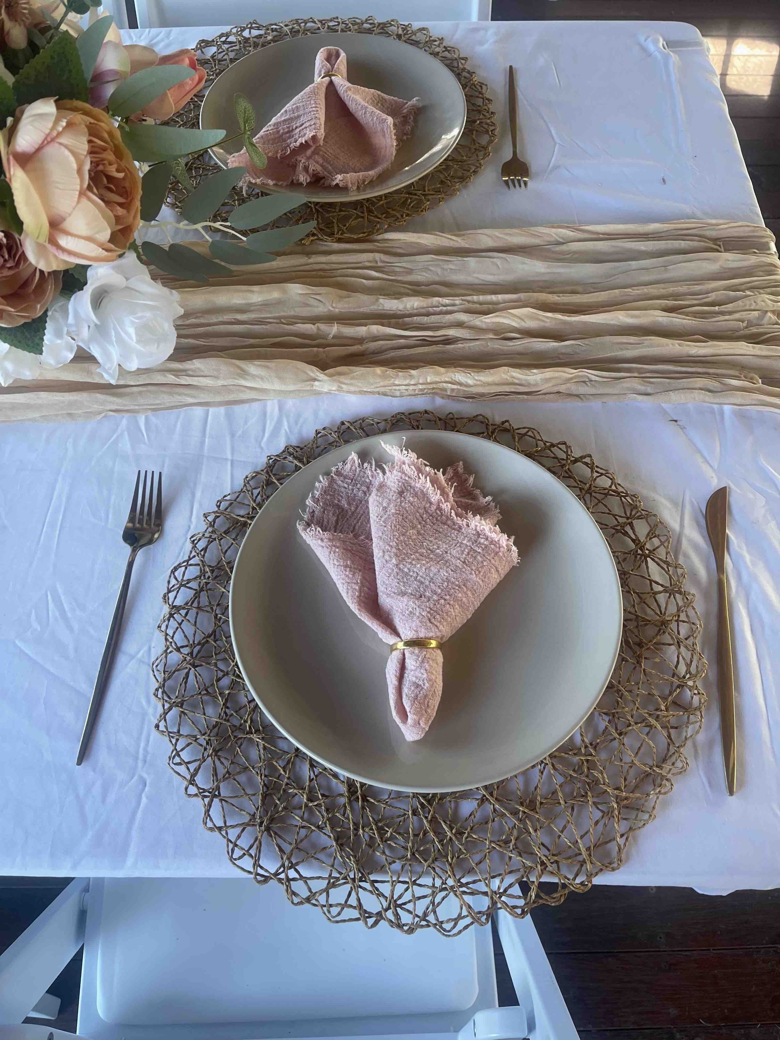 A dining table set for a meal with a white plate, pink napkin, fork, and knife. There is a woven placemat underneath the plate, a floral centerpiece on the side, and a beige table runner. The table is covered with a white tablecloth.