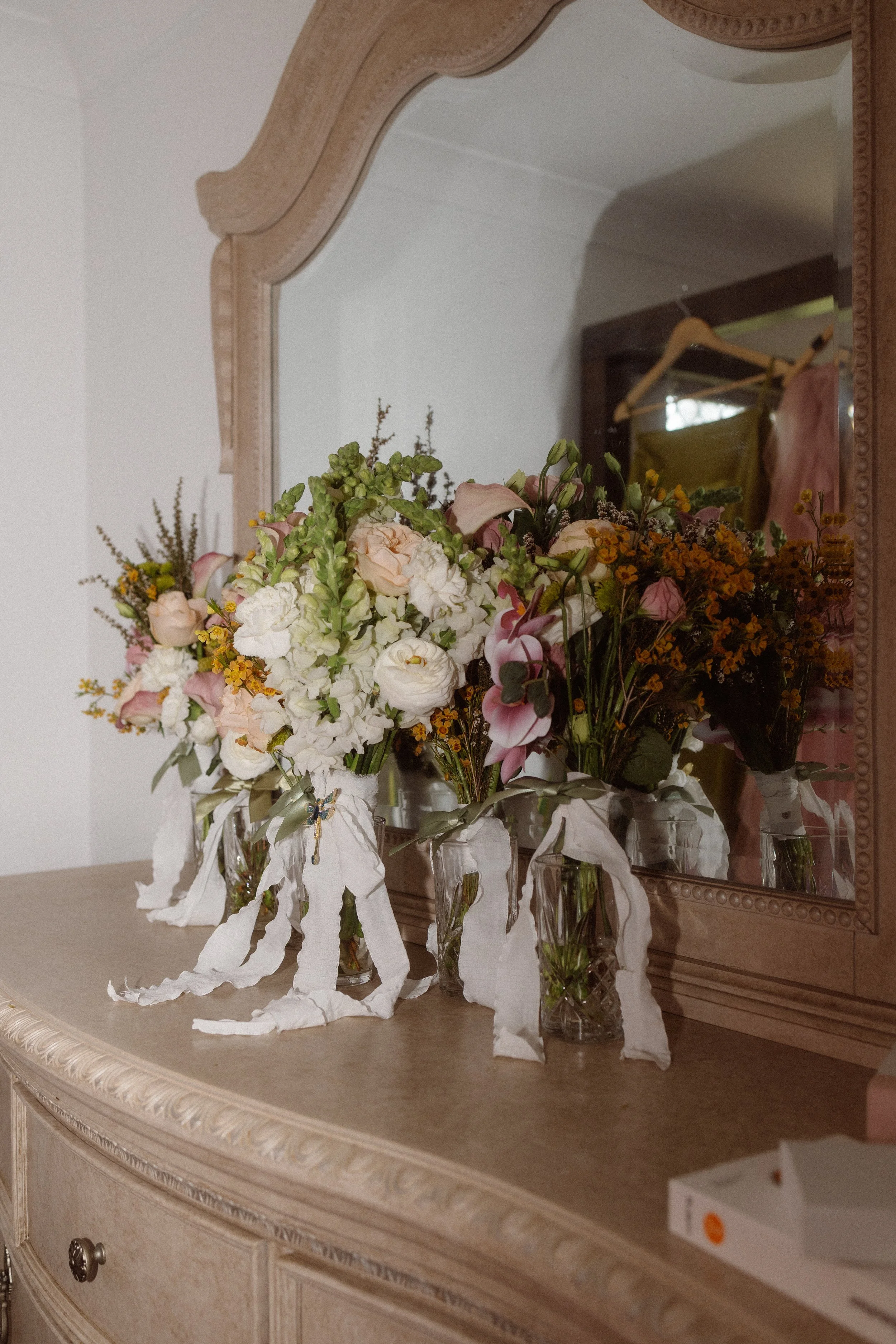Four glass vases with white and pink flowers on a wooden dresser, reflected in a large mirror with a decorative frame, with clothing hanging in the background.