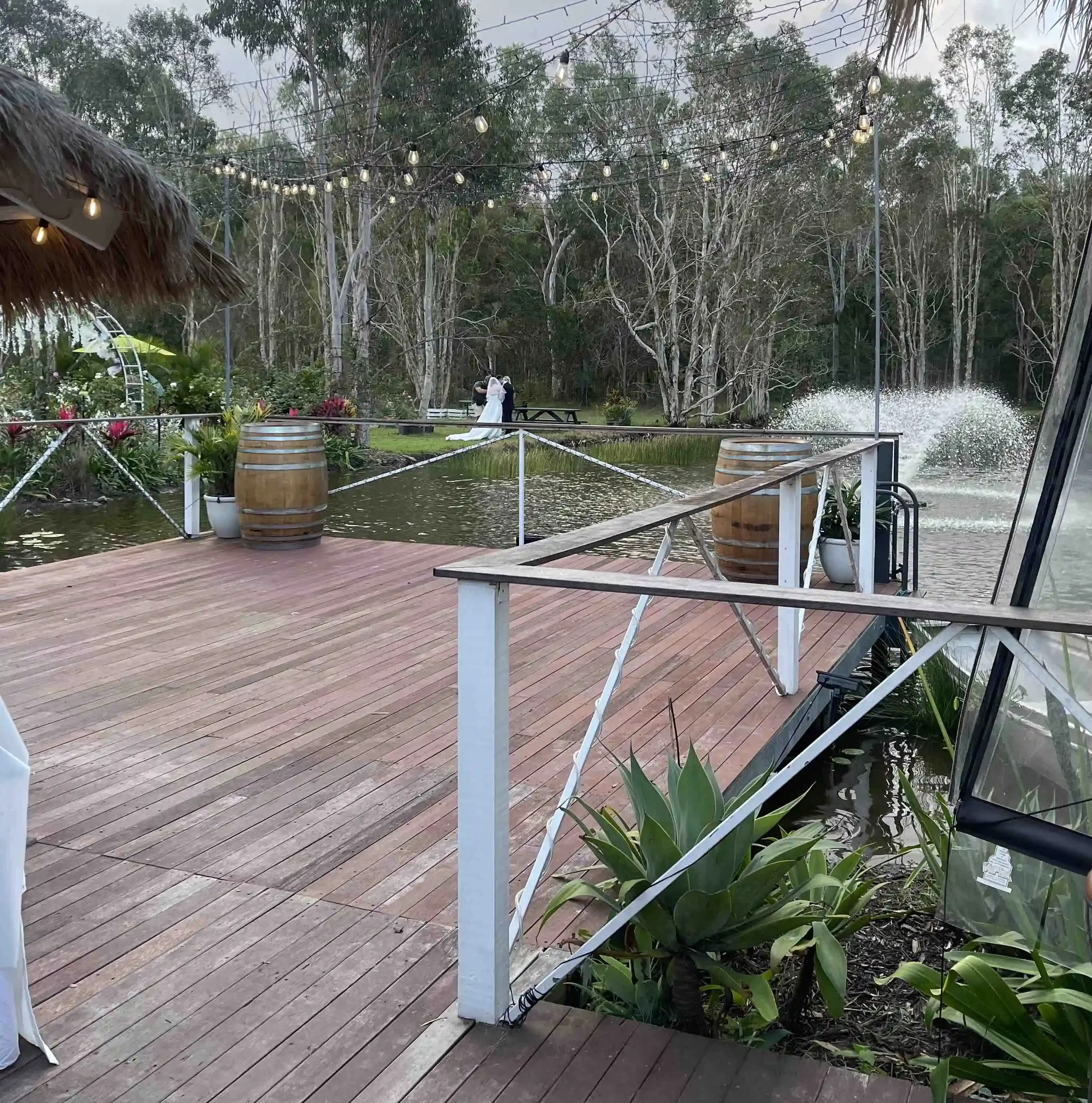 Outdoor scene with string lights hanging overhead, trees in the background, a thatched roof structure on the left, potted plants, a wooden deck, and a couple of people near a pond or water feature with a fountain.