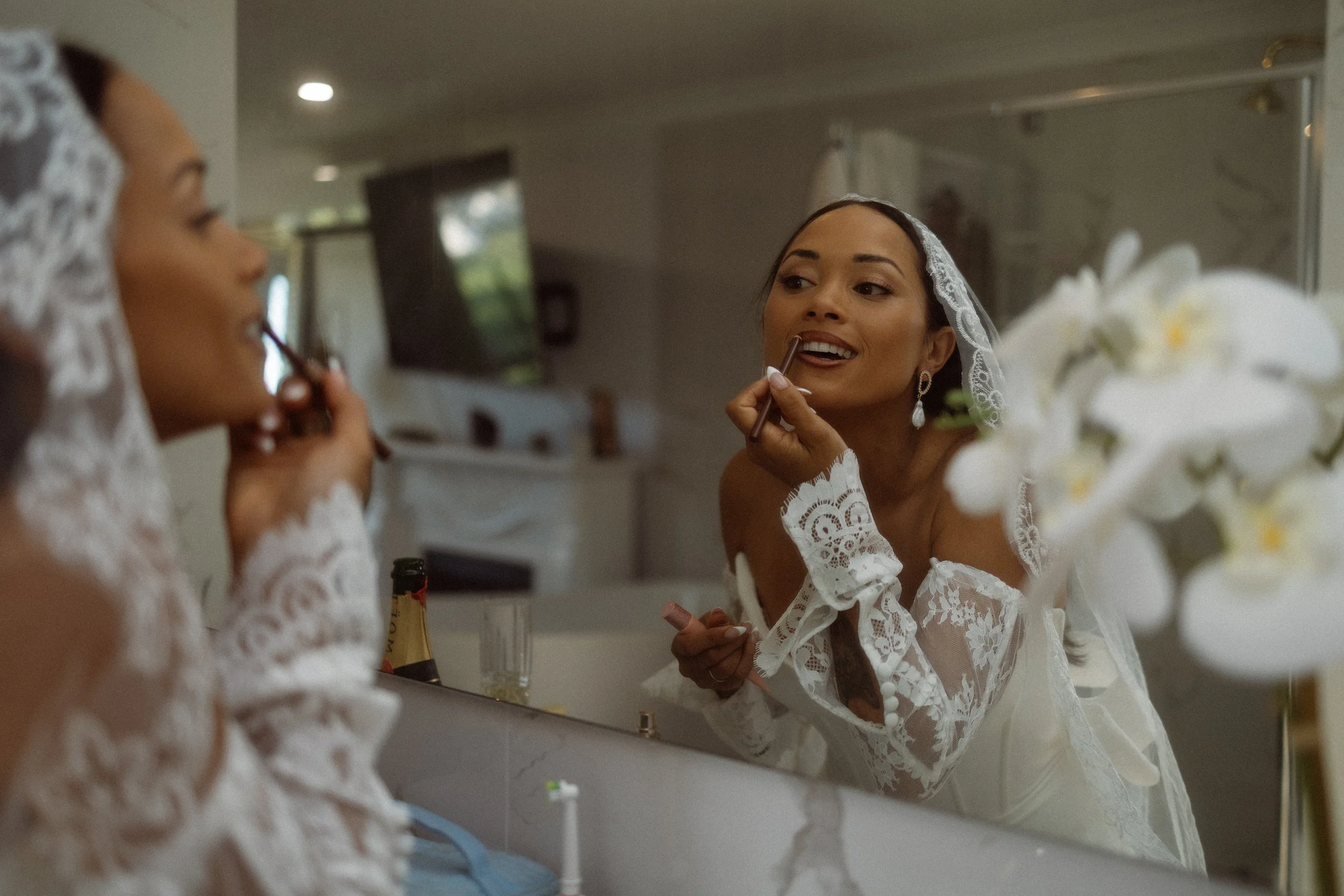 A bride in a white lace wedding dress and veil applies lipstick in front of a mirror in a bathroom with flowers and toiletries visible.