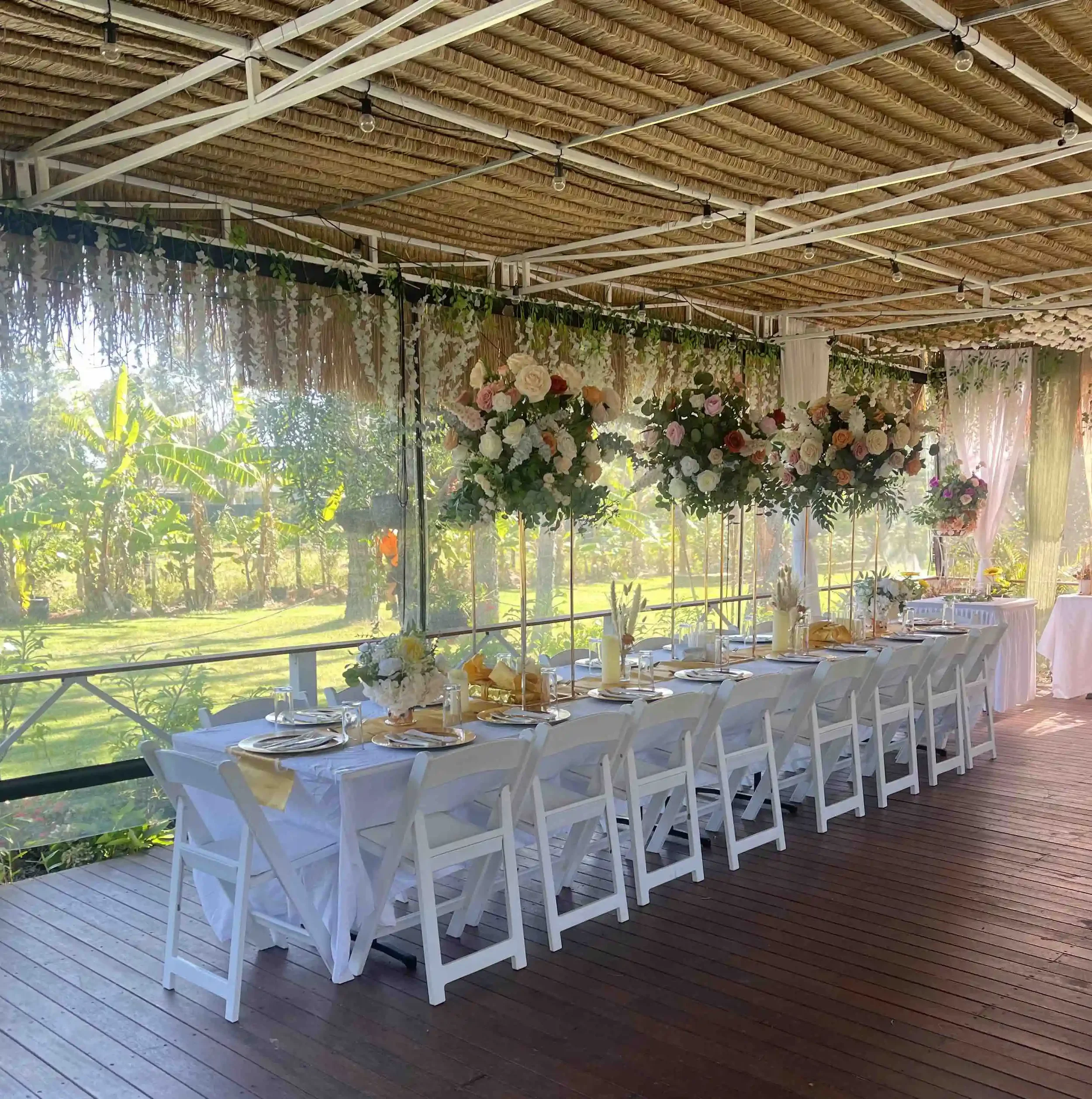 A long banquet table set for an event with white chairs, floral centerpieces, candles, and tableware, located outdoors under a thatched roof with string lights and greenery.