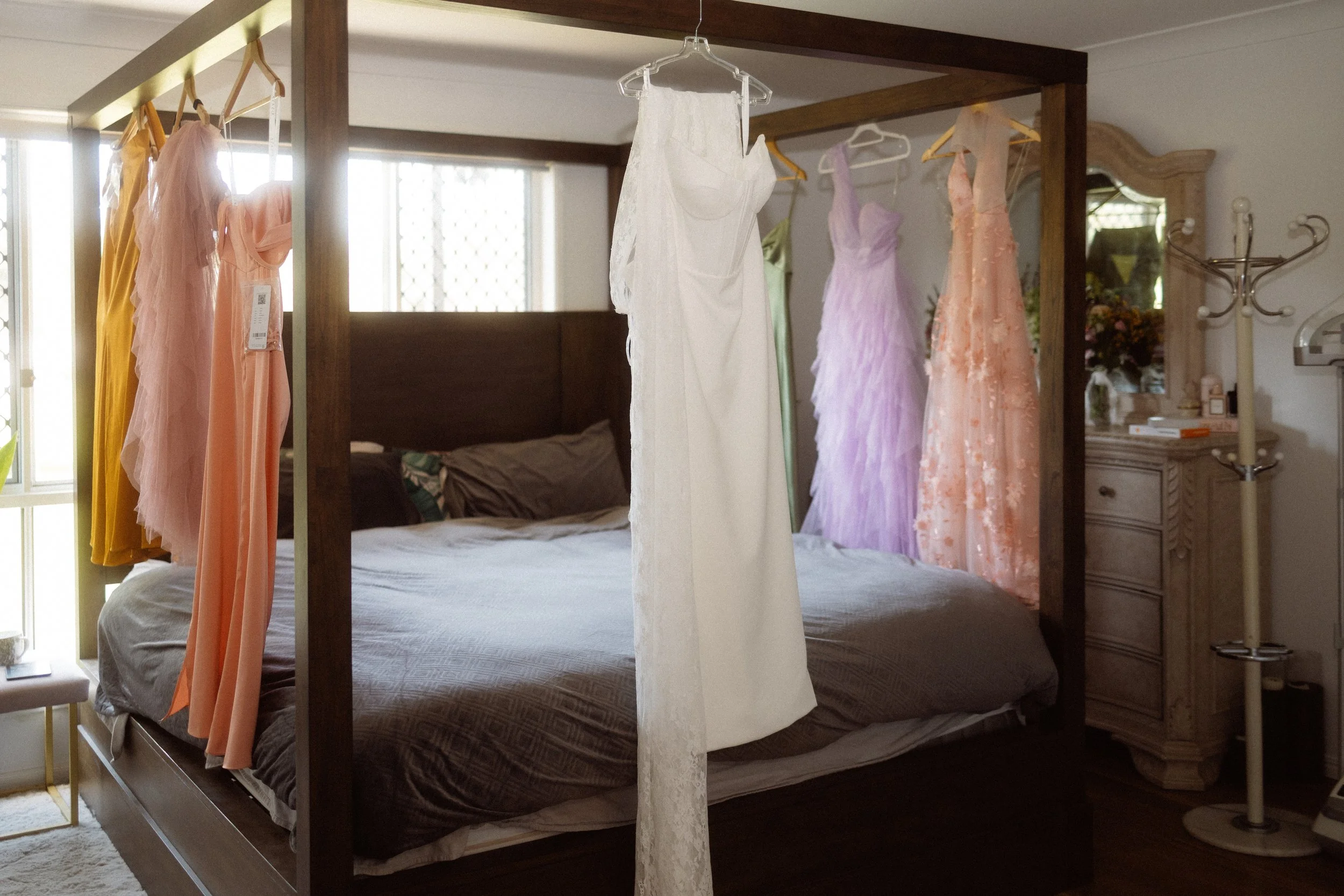 A bedroom with a four-poster bed and a wooden frame, featuring various dresses hanging on the side and behind the bed, including a white wedding gown in the center, pastel-colored and peach dresses, with windows letting in natural light.