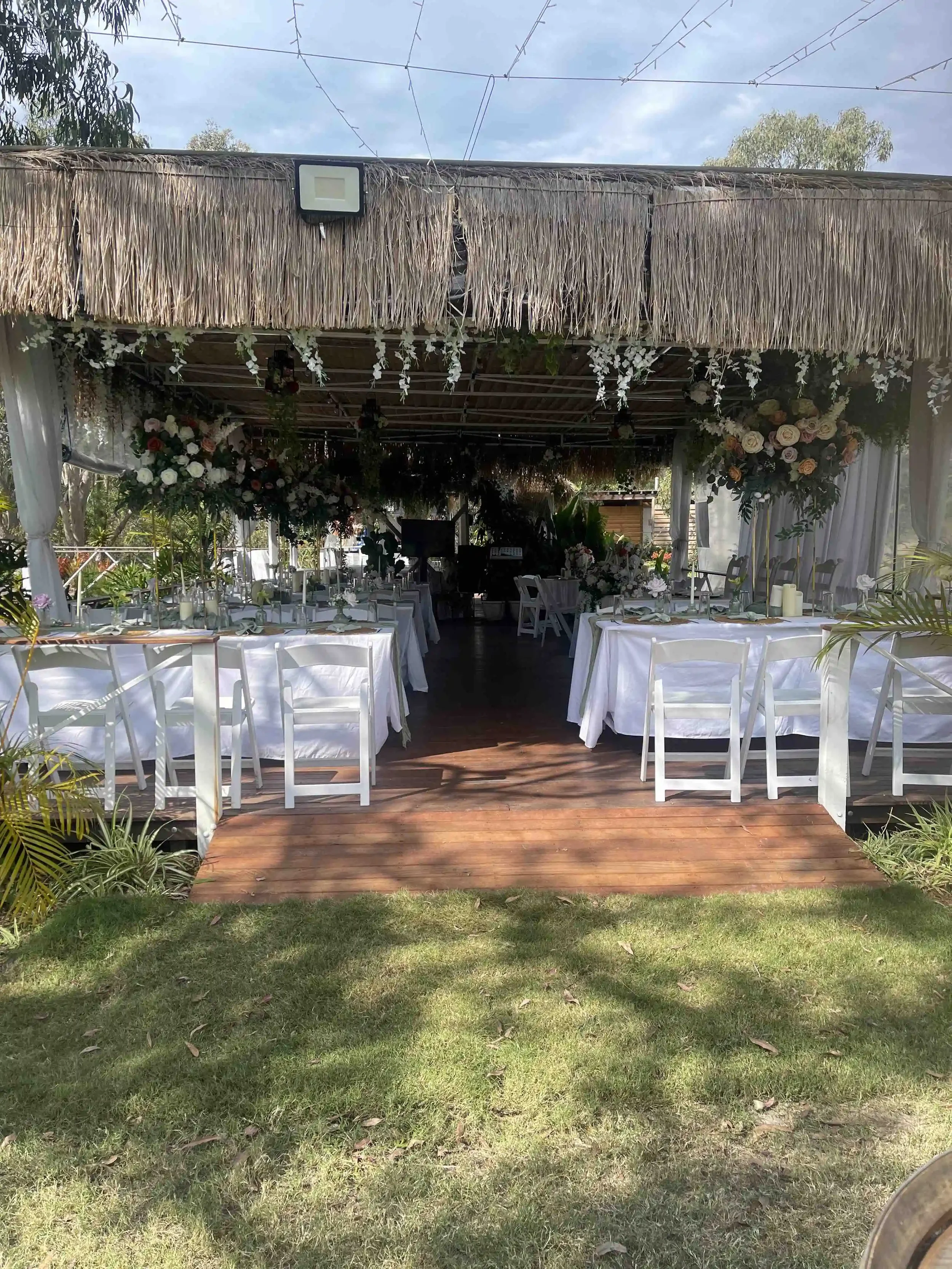 An outdoor event setup with white tables and chairs under a thatched roof, decorated with floral arrangements, surrounded by greenery and trees.
