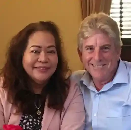 A woman and a man smiling and sitting together in a warmly lit room with a window and curtains behind them.