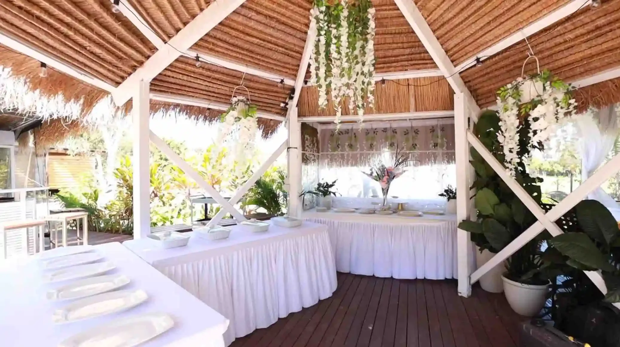 Beachside outdoor dining area with white tablecloths, plates, and plants, under a thatched roof with hanging white flowers and greenery.