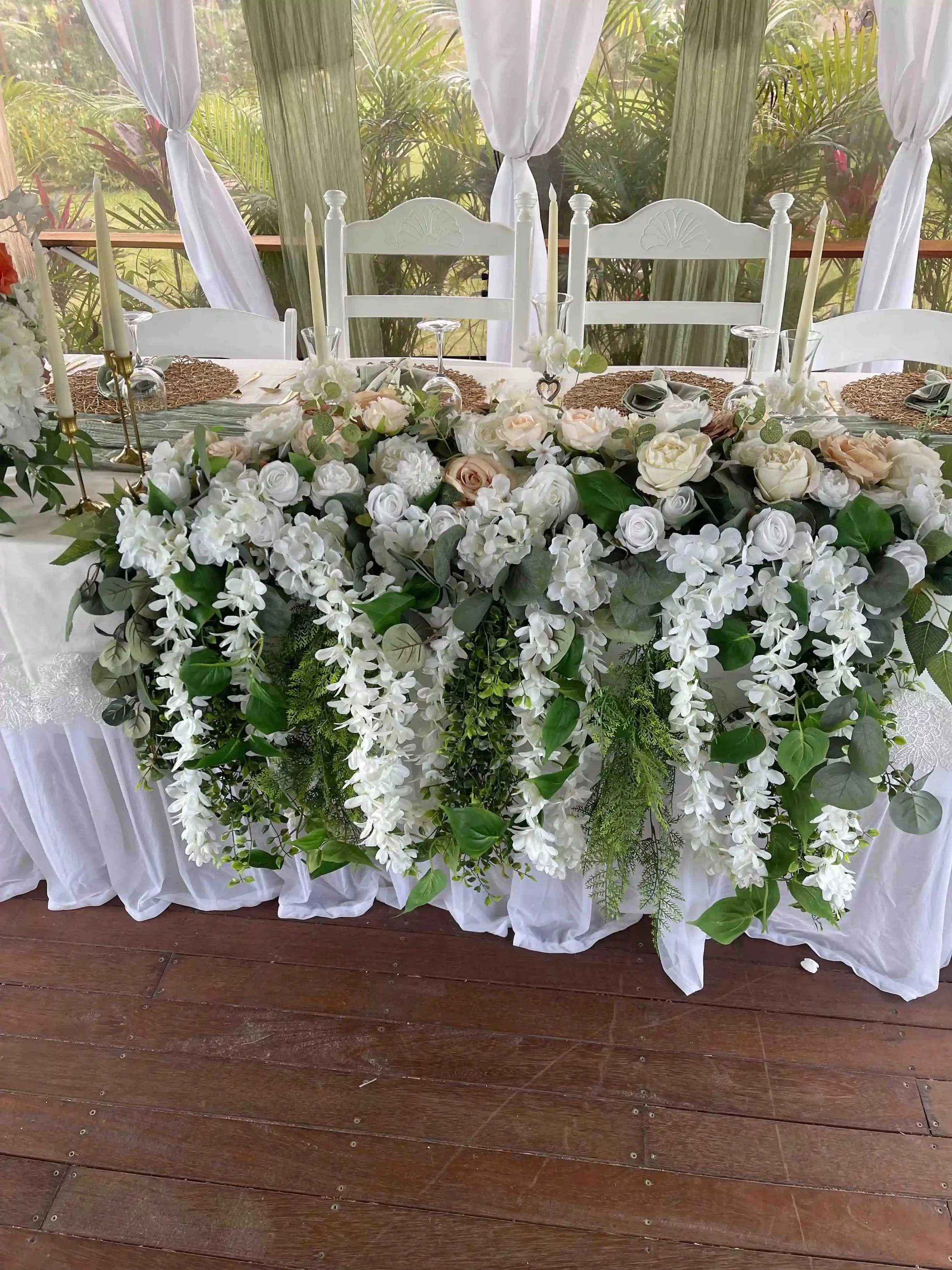 A decorated table with a floral arrangement featuring white and blush roses, greenery, and cascading white flowers. The table has white chairs with a sheer white curtain backdrop, set for a special occasion or celebration.