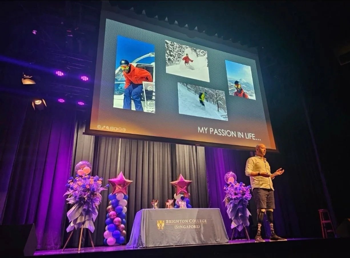 A man with a prosthetic leg giving a presentation on a stage decorated with purple and pink balloons and flowers, with a large screen displaying photos of skiing activities and the text 'My Passion in Life'.