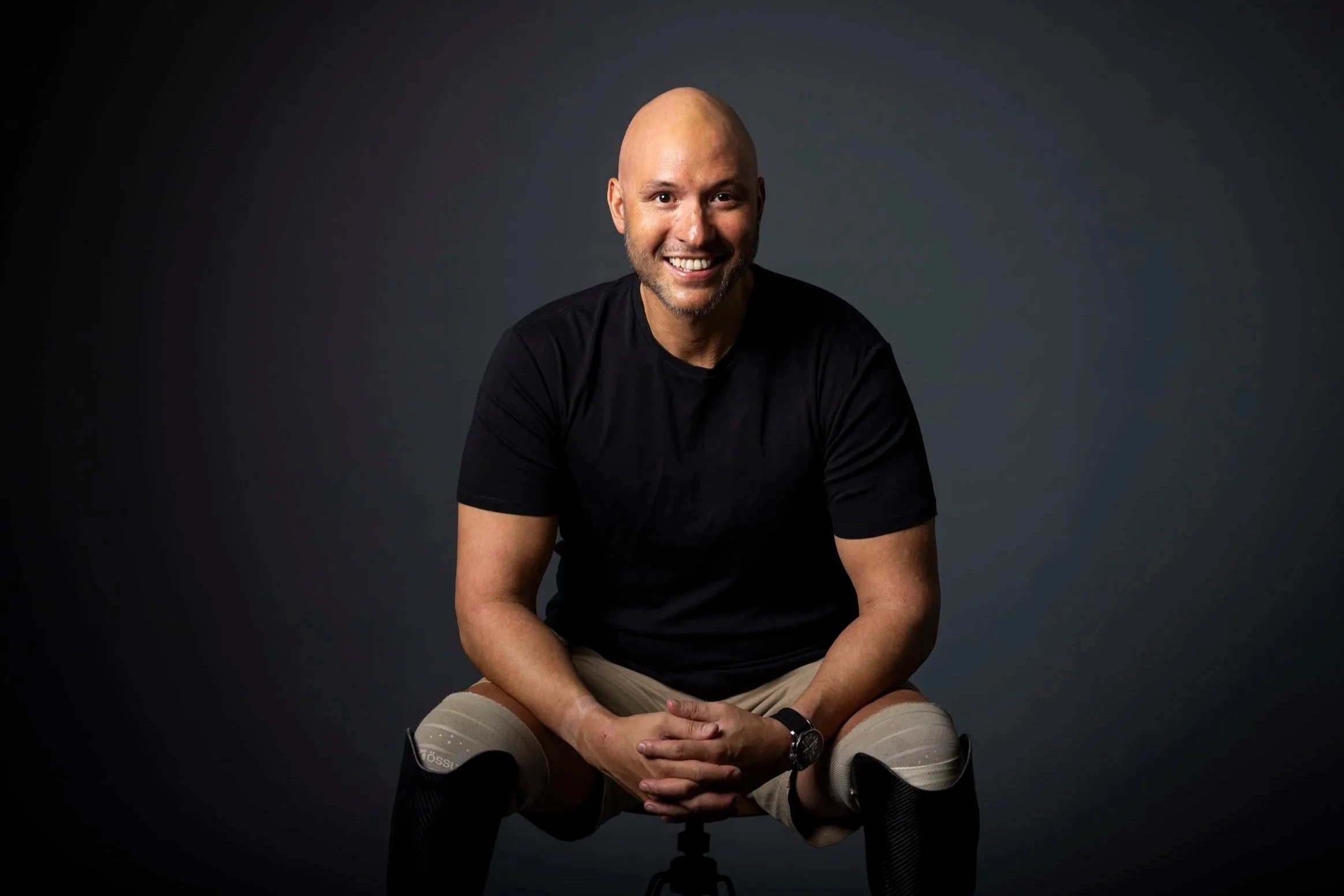 A smiling man with a bald head and beard, wearing a black T-shirt, beige shorts, and a watch, sitting on a chair against a dark gray background.
