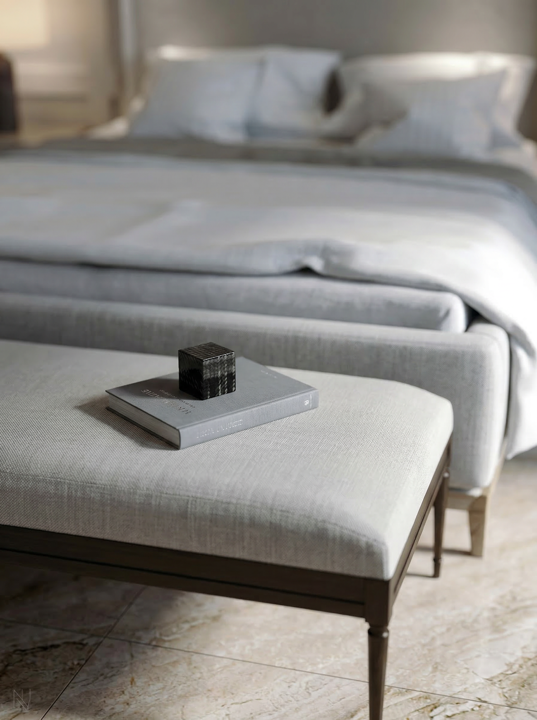 A bedroom with a bed in the background, a gray upholstered bench at the foot of the bed, a hardcover book, and a small decorative black cube on the bench.
