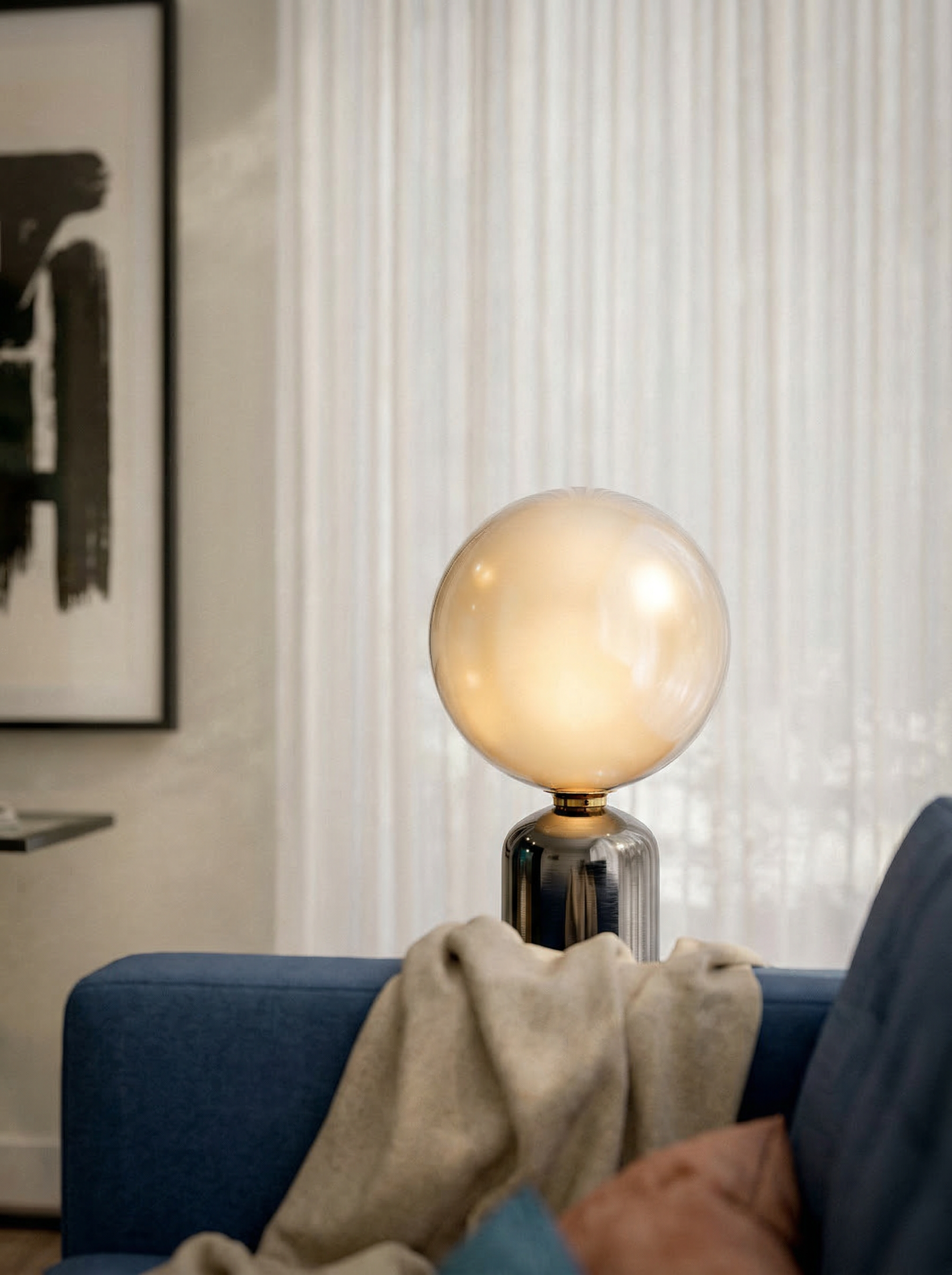 A modern table lamp with a glowing spherical frosted glass shade and a reflective metallic base, placed on a side table in a contemporary living room.