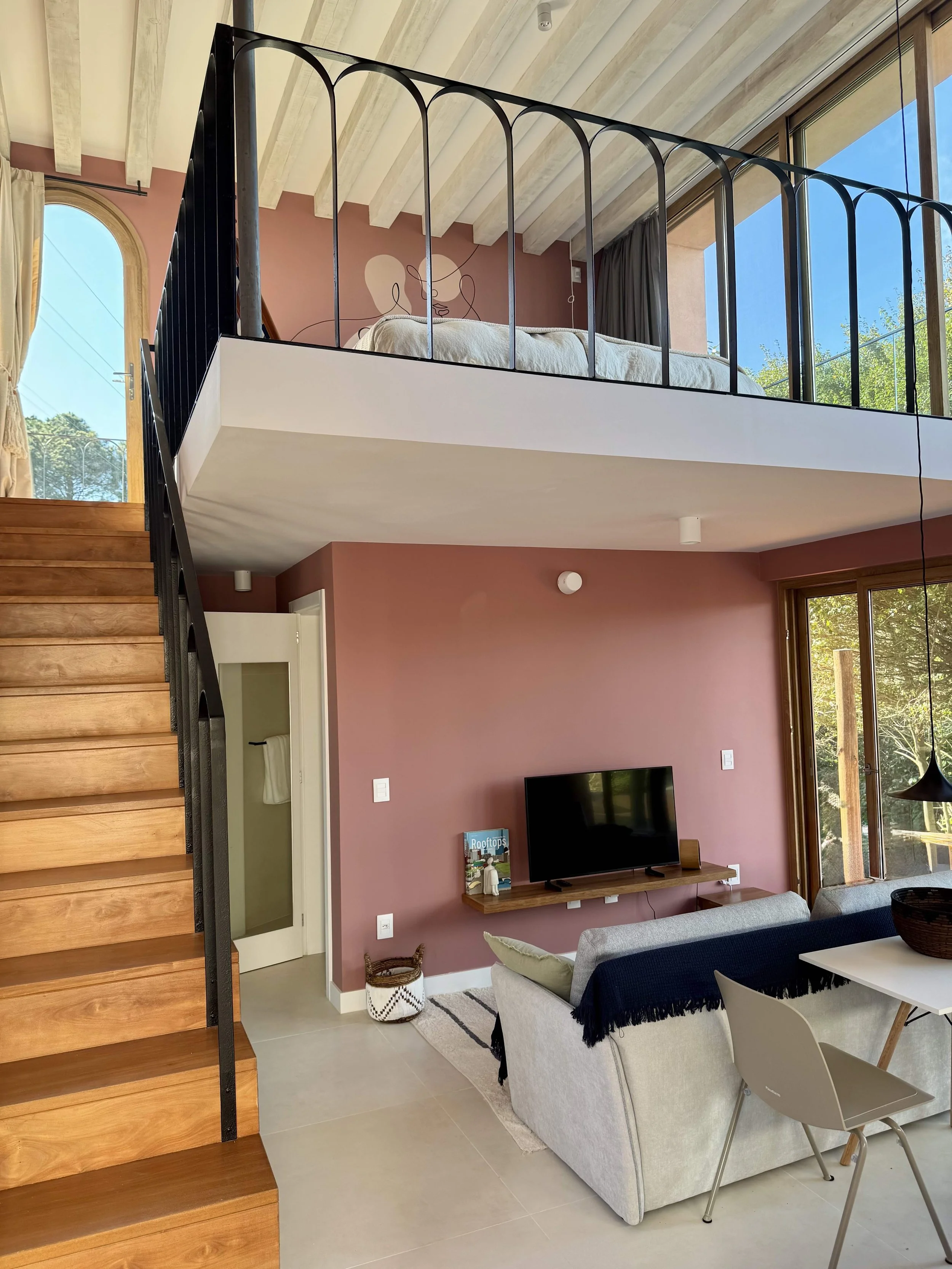 Open-plan living room with a sofa, coffee table, and wall-mounted TV on a pink accent wall, with large windows and a loft bedroom above, connected by a wooden staircase.