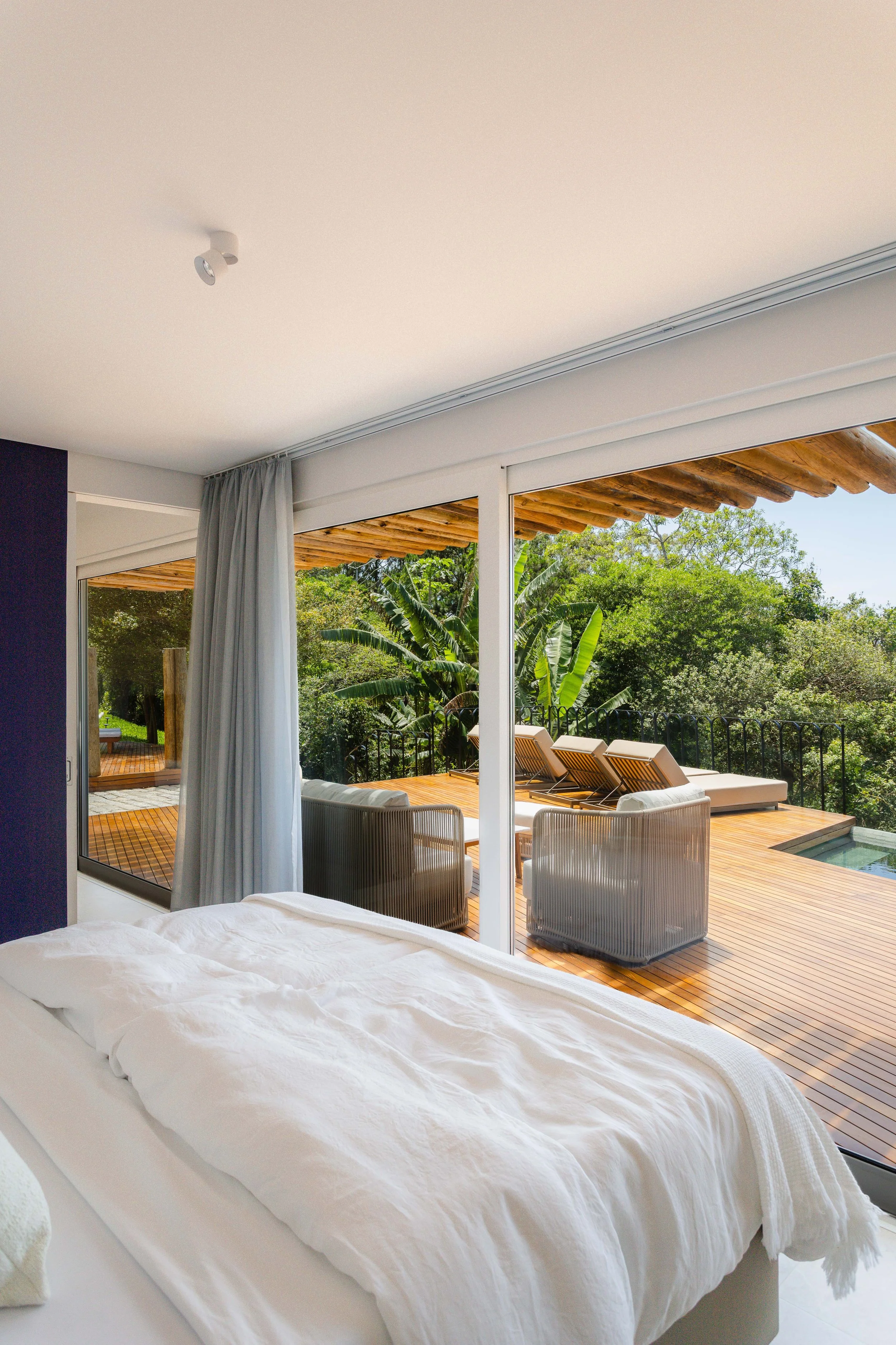 Bedroom with a large glass sliding door leading to a outdoor patio with lounge chairs and lush greenery.