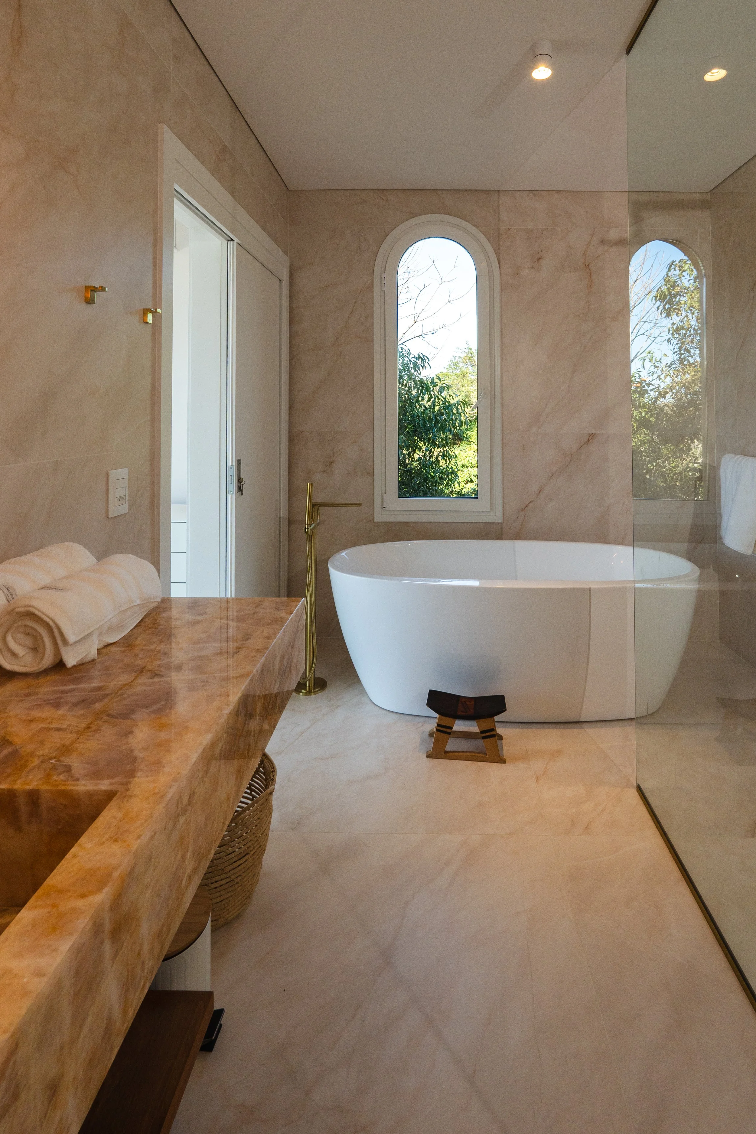 Modern bathroom with a large white oval bathtub, a window with trees outside, a marble countertop, and a glass shower enclosure.