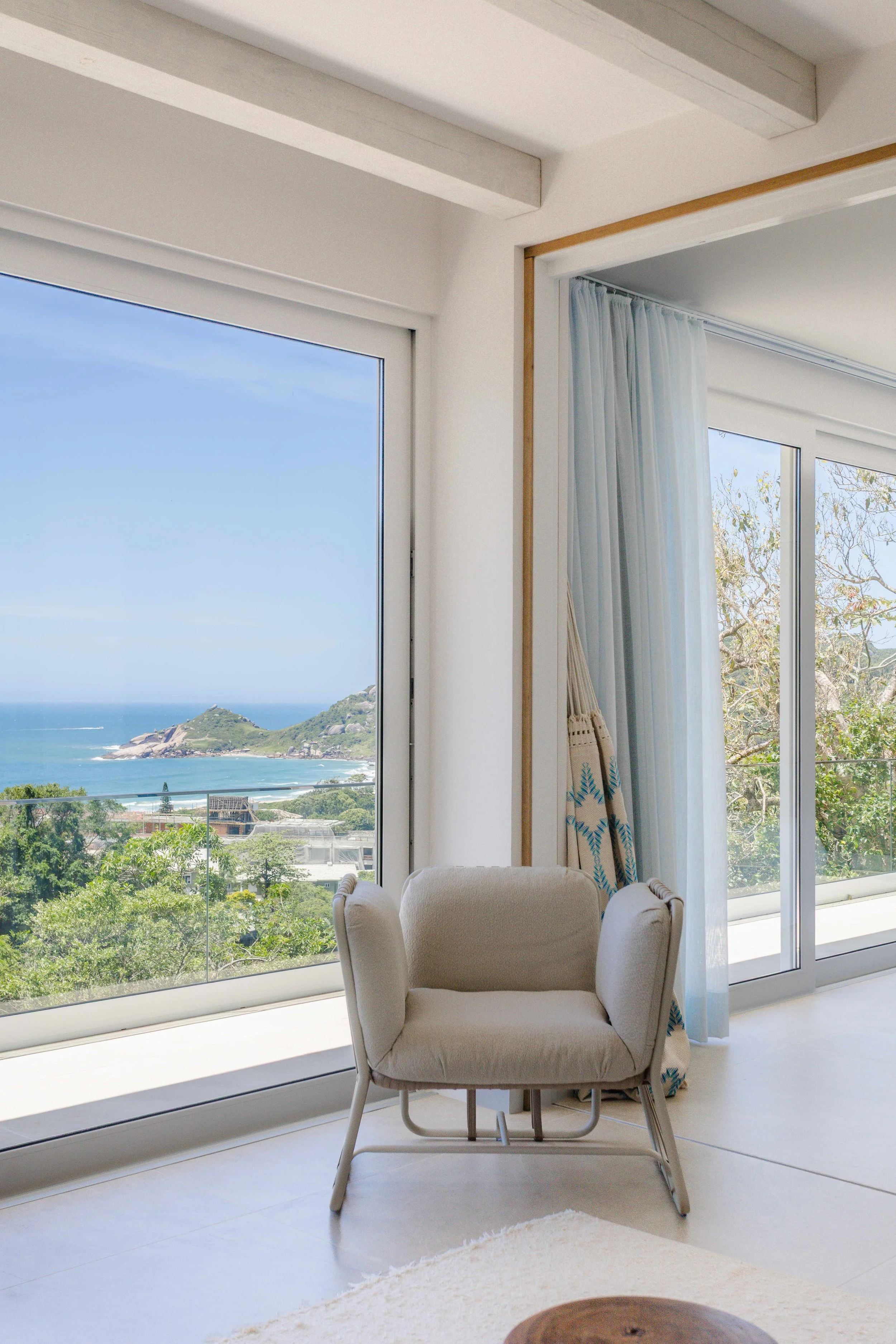 Living room with large glass windows overlooking a coastal landscape with hills, ocean, and sky; beige armchair near sliding glass door with sheer curtains.