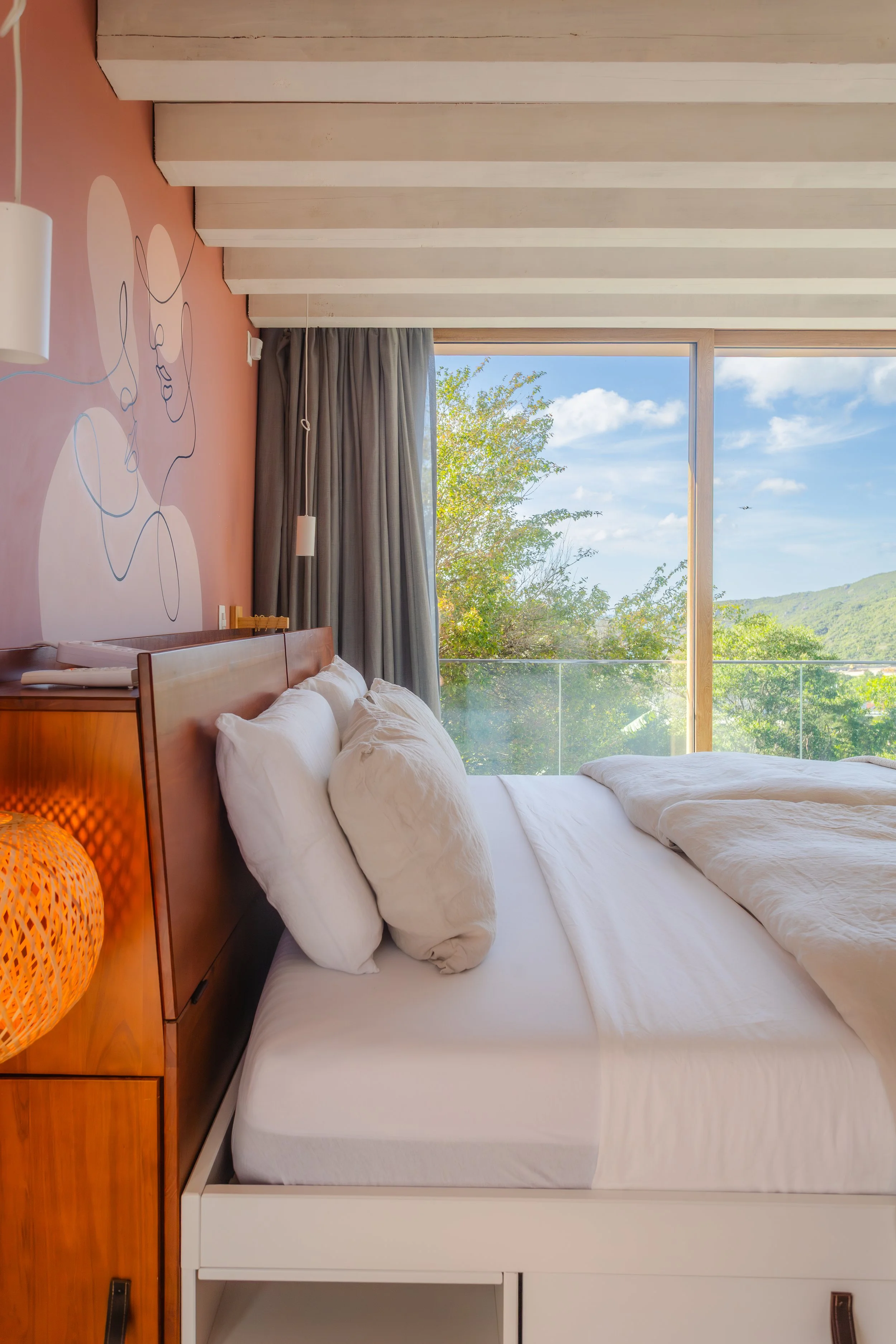 A cozy bedroom with a bed, white pillows, and a view of green trees and a blue sky through large glass windows.