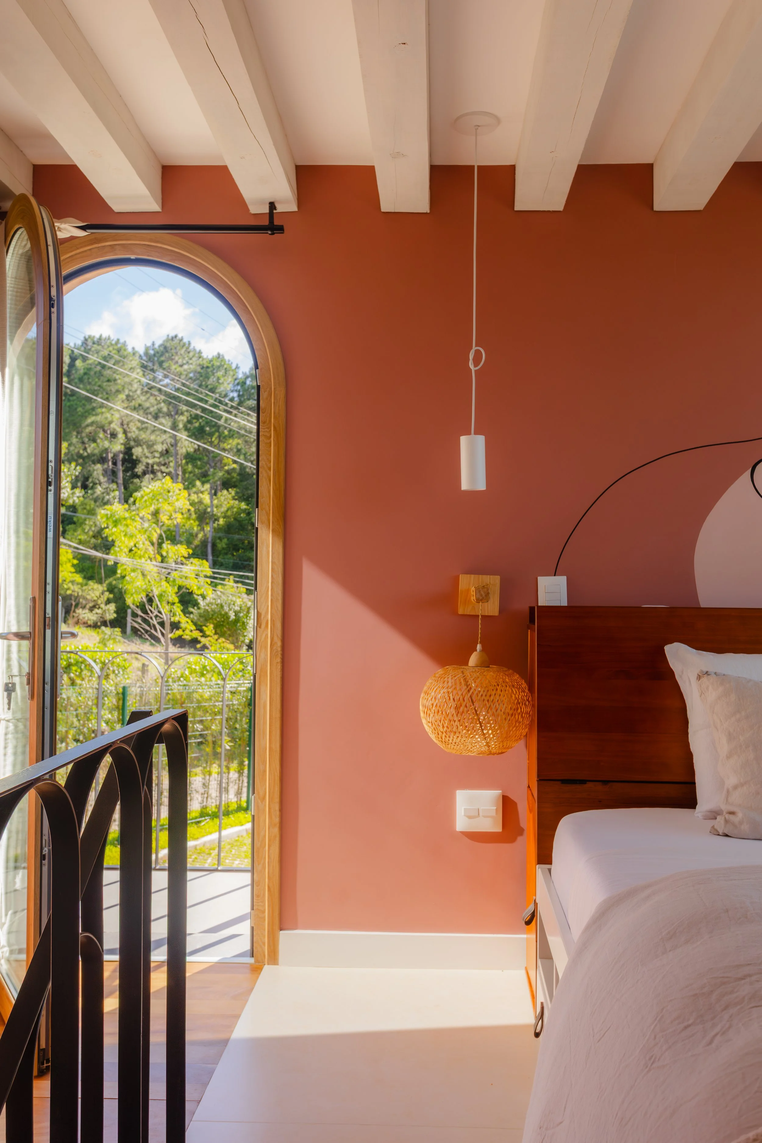 Bright bedroom with pink wall, featuring a wooden bed, a hanging white pendant light, a woven basket lamp, and an arched door with a view of trees outside.