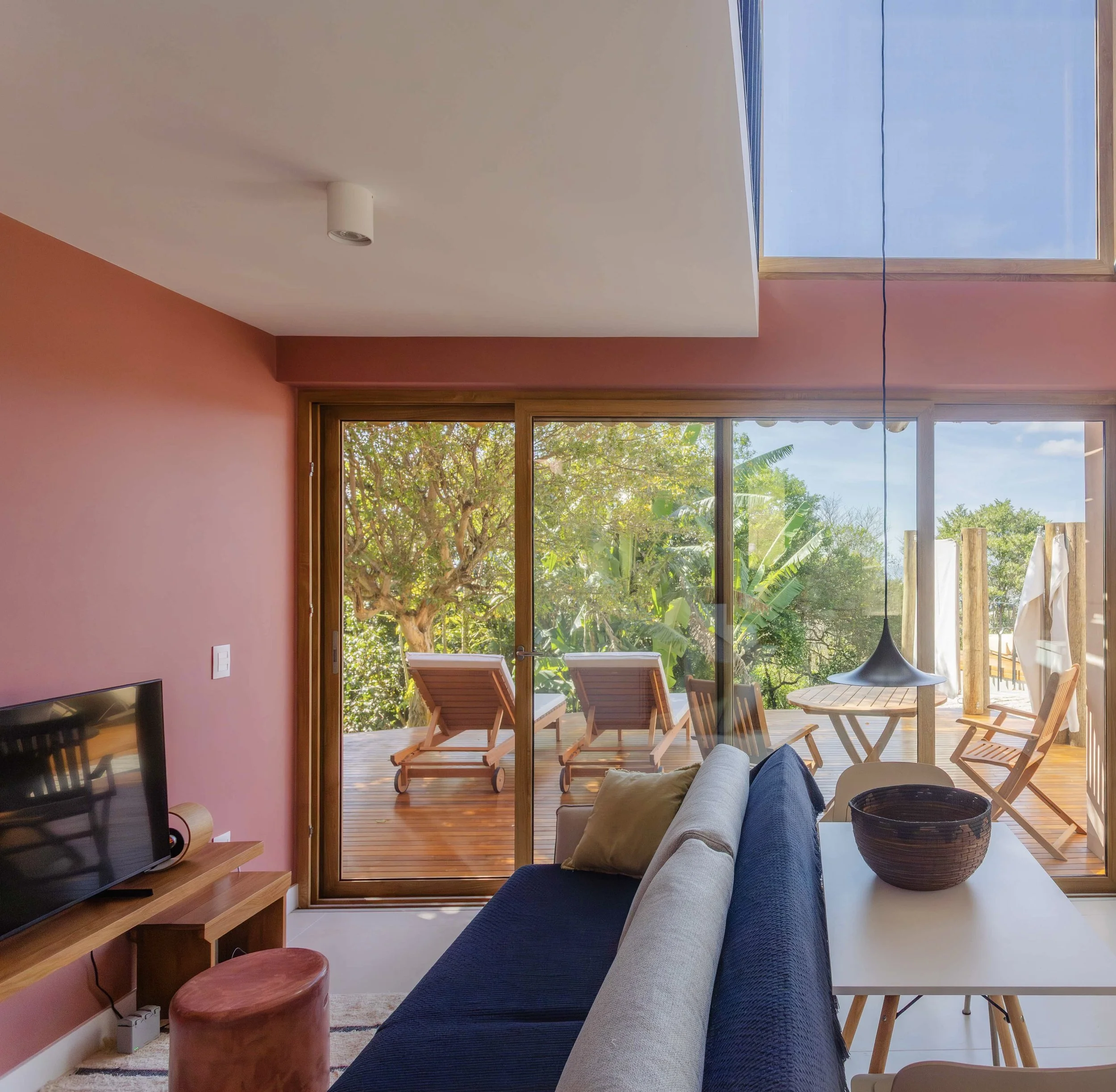 Living room with pink wall, sliding glass door leading to outdoor deck with two lounge chairs, trees outside, and a round table with four chairs inside, along with ceiling window and ceiling light.