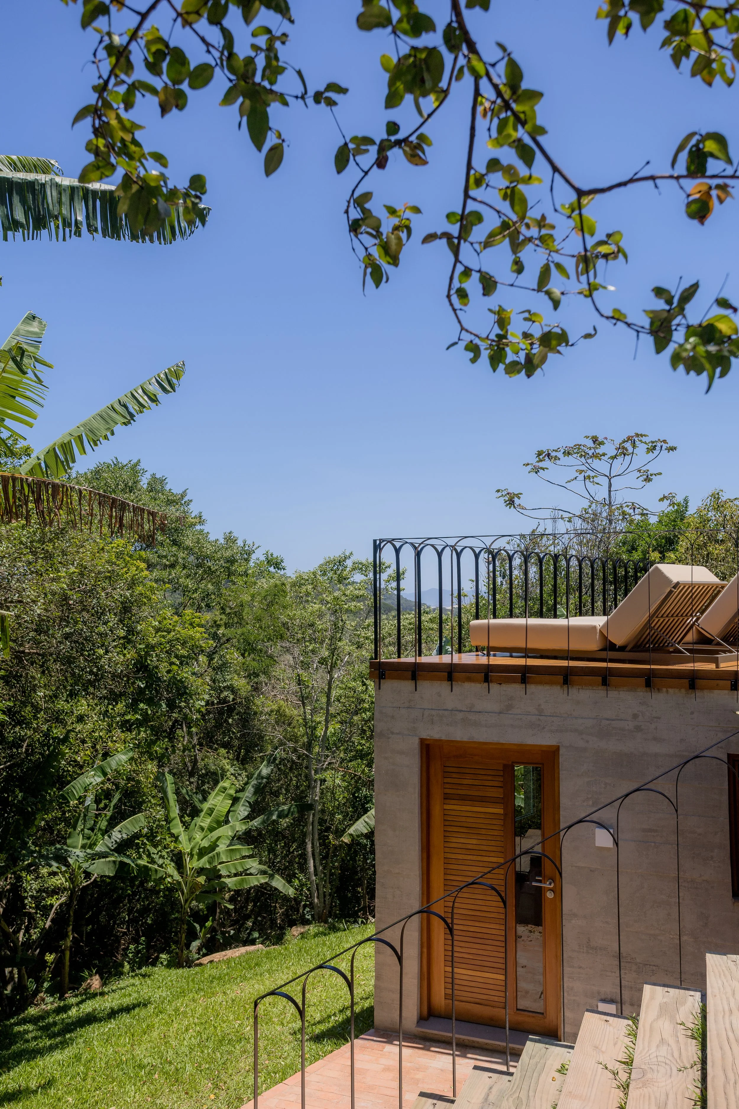 A house with a wooden door and a balcony with lounge chairs on top, surrounded by lush green trees and plants, under a clear blue sky.
