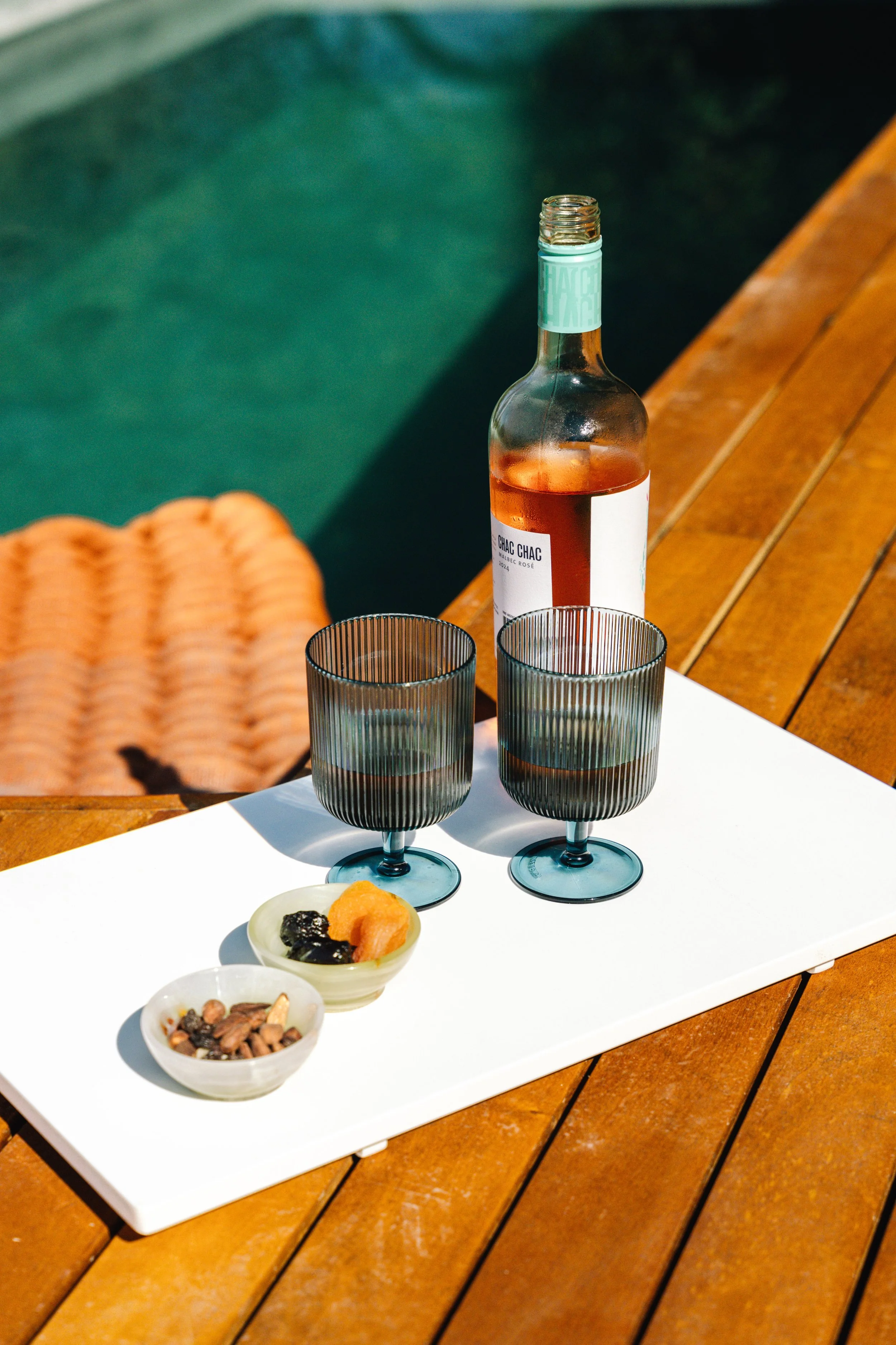 A bottle of rosé wine and two empty glasses on a white tray, with small bowls of dried fruits and nuts, on a wooden table outdoors.