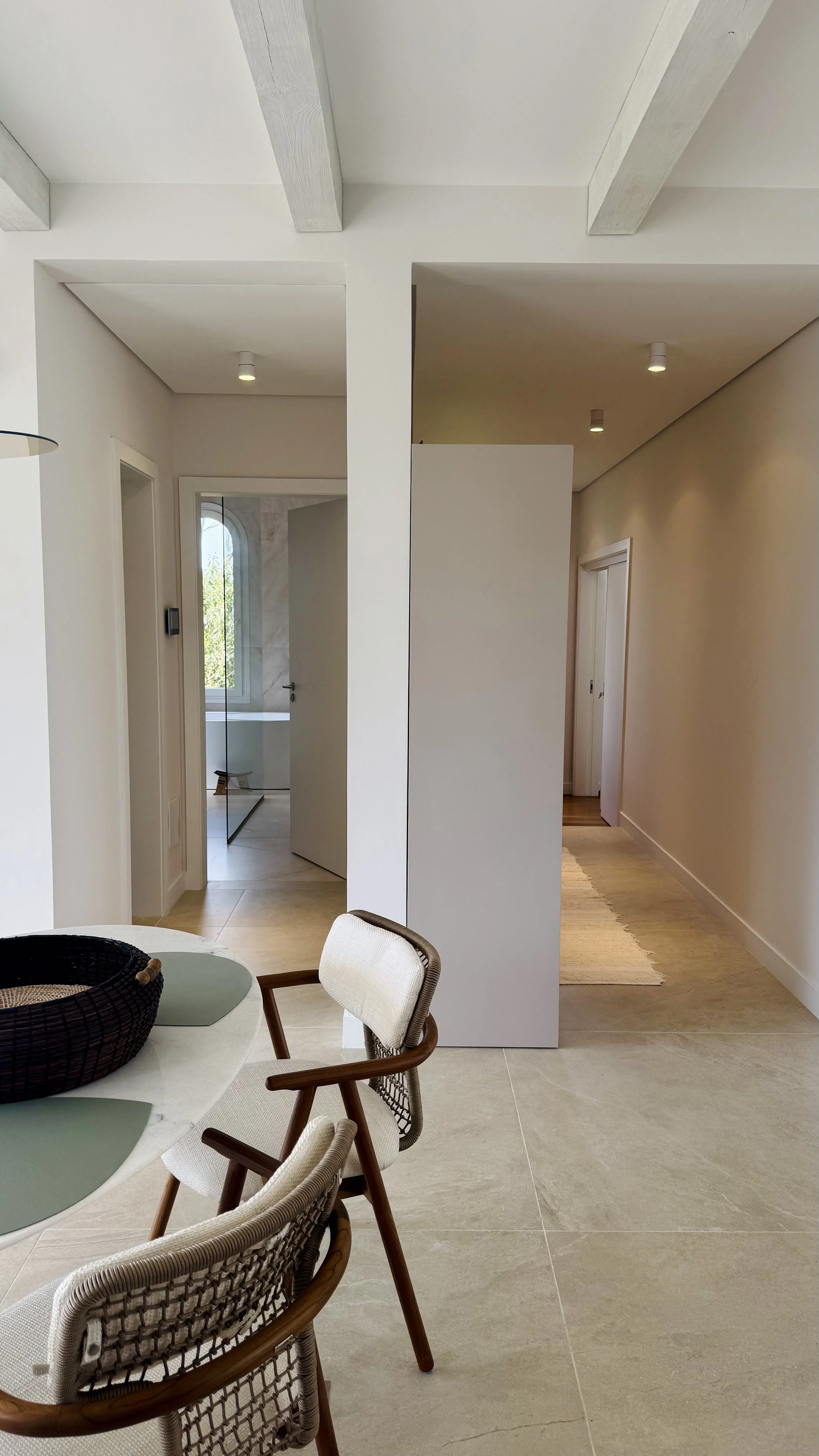 Interior view of a modern, minimalist home with a dining area in the foreground, featuring beige chairs and a round table. In the background, a corridor splits into two paths with white walls and ceiling beams, leading to other rooms with natural light coming through the windows.