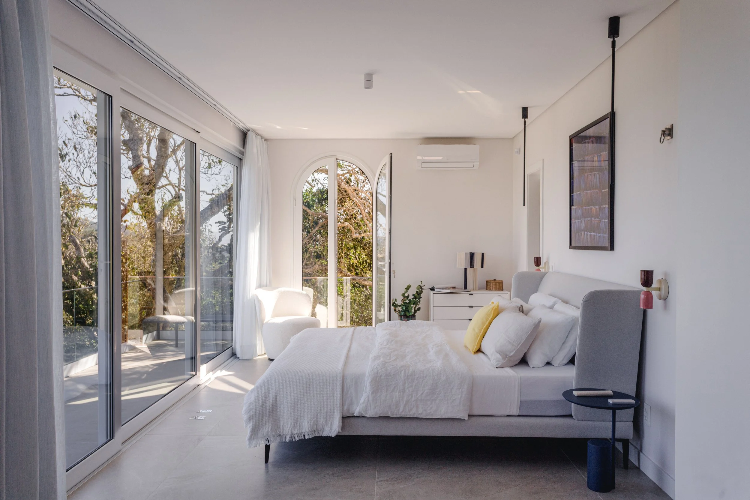 Bright bedroom with large glass sliding doors and balcony, white bedding, yellow pillow, nightstand with lamp, artwork on white wall, and minimal decor.