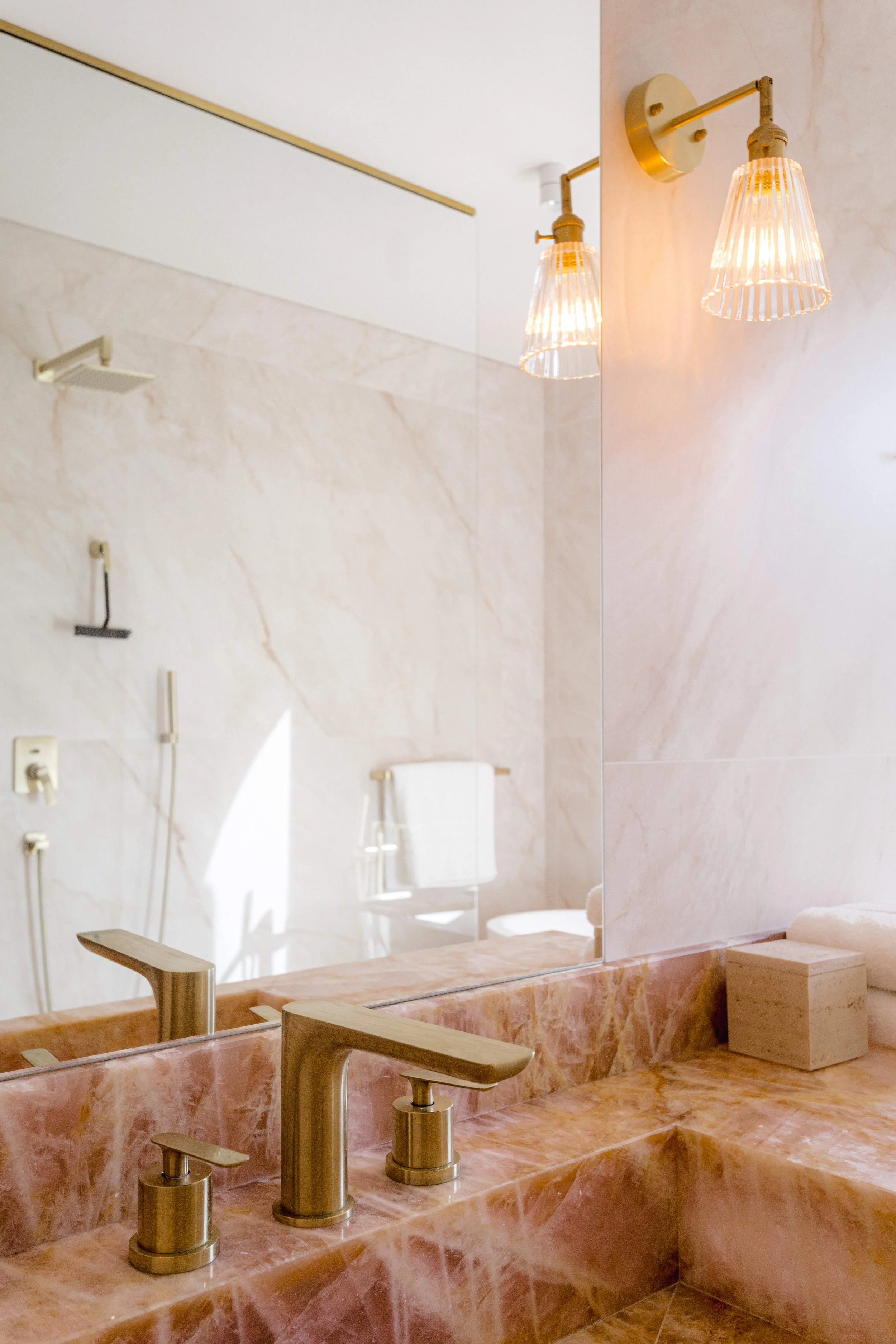 A bathroom vanity with a rose-colored marble countertop, a brass faucet, a large mirror, two wall-mounted brass light fixtures, a boxed tissue, and a towel rack reflected in the mirror.