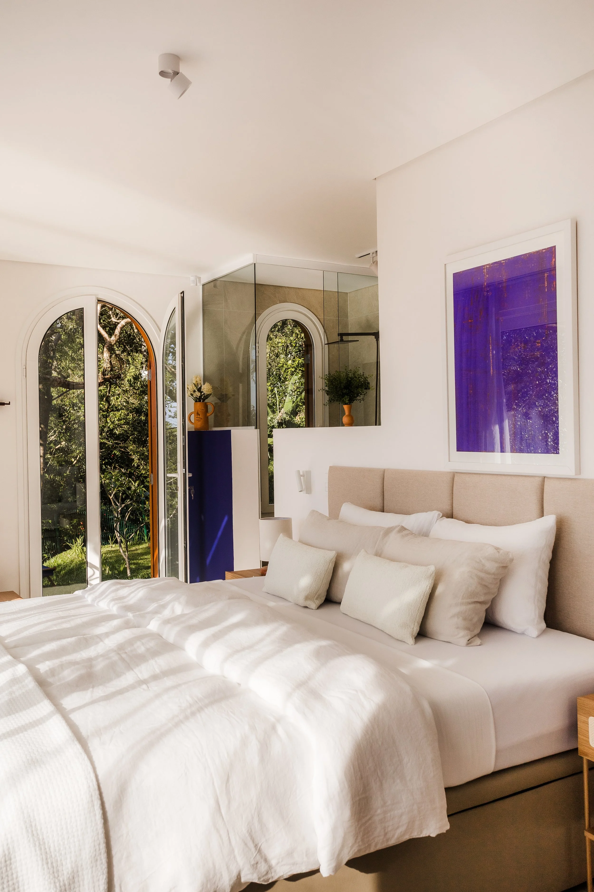 Bright, modern bedroom with a large bed, beige headboard, multiple white and beige pillows, light-colored bedding, and a framed purple artwork above the bed. French doors with glass panels open to a garden with trees outside, and a glass-walled bathroom area with a shower is visible in the background.