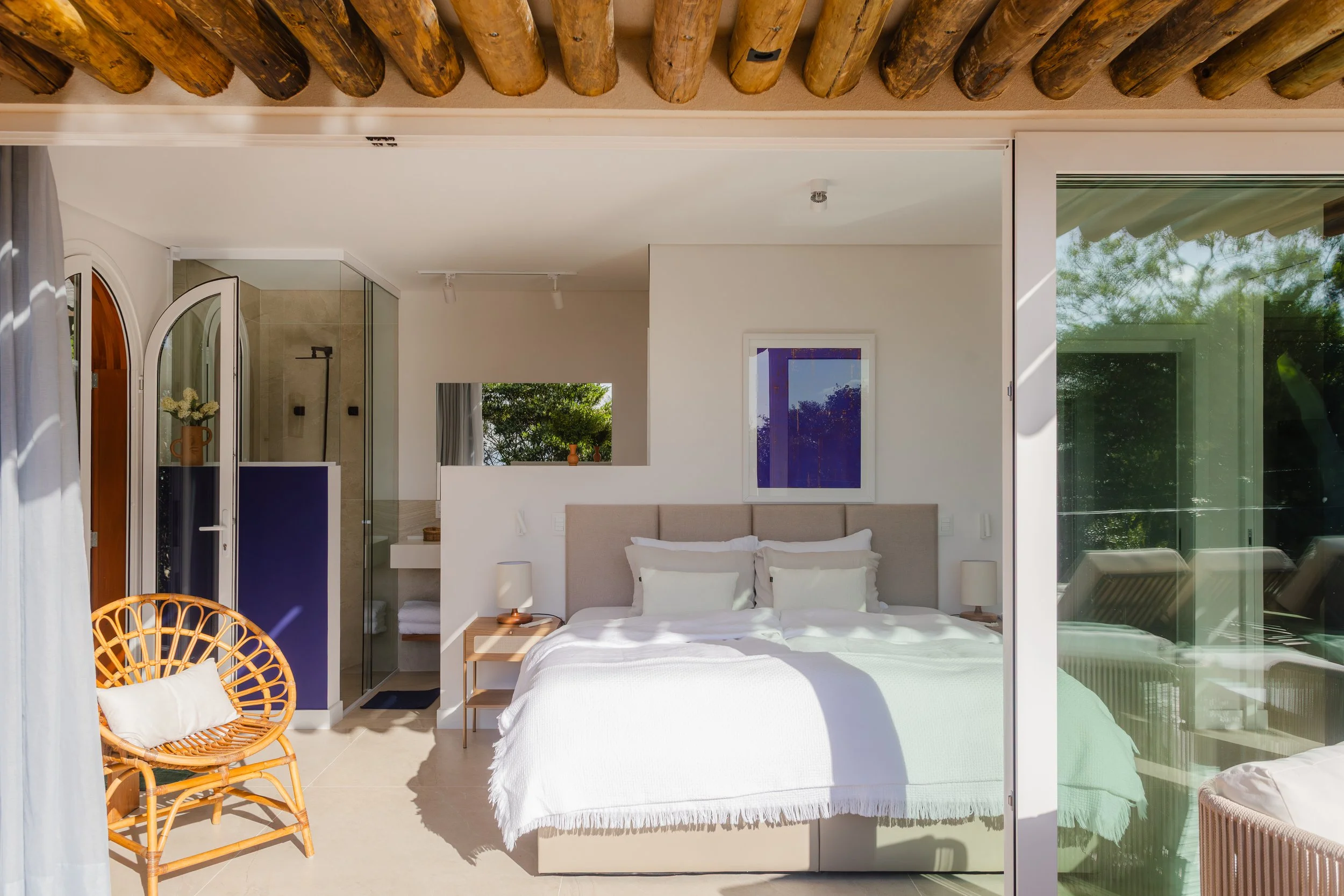 A bedroom with a large bed, beige upholstered headboard, and white bedding. Two white nightstands with lamps on each side of the bed. An open sliding glass door on the right leading to an outdoor area with greenery. A small window above the headboard. A wicker chair with a white cushion on the left. An alcove with a door leading to a bathroom with a shower visible. Wooden beams on the ceiling.