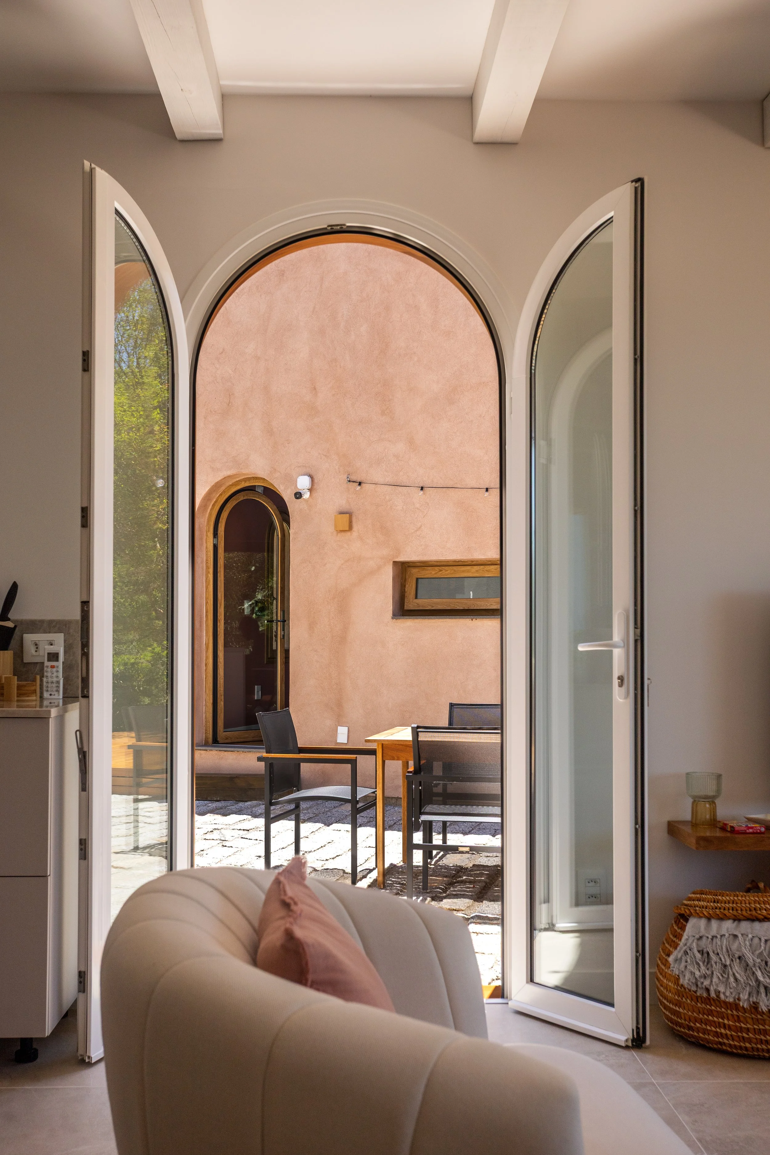 Open glass doors leading to an outdoor patio with black chairs, a wooden table, and a pink stucco wall with an arched window and doorway.