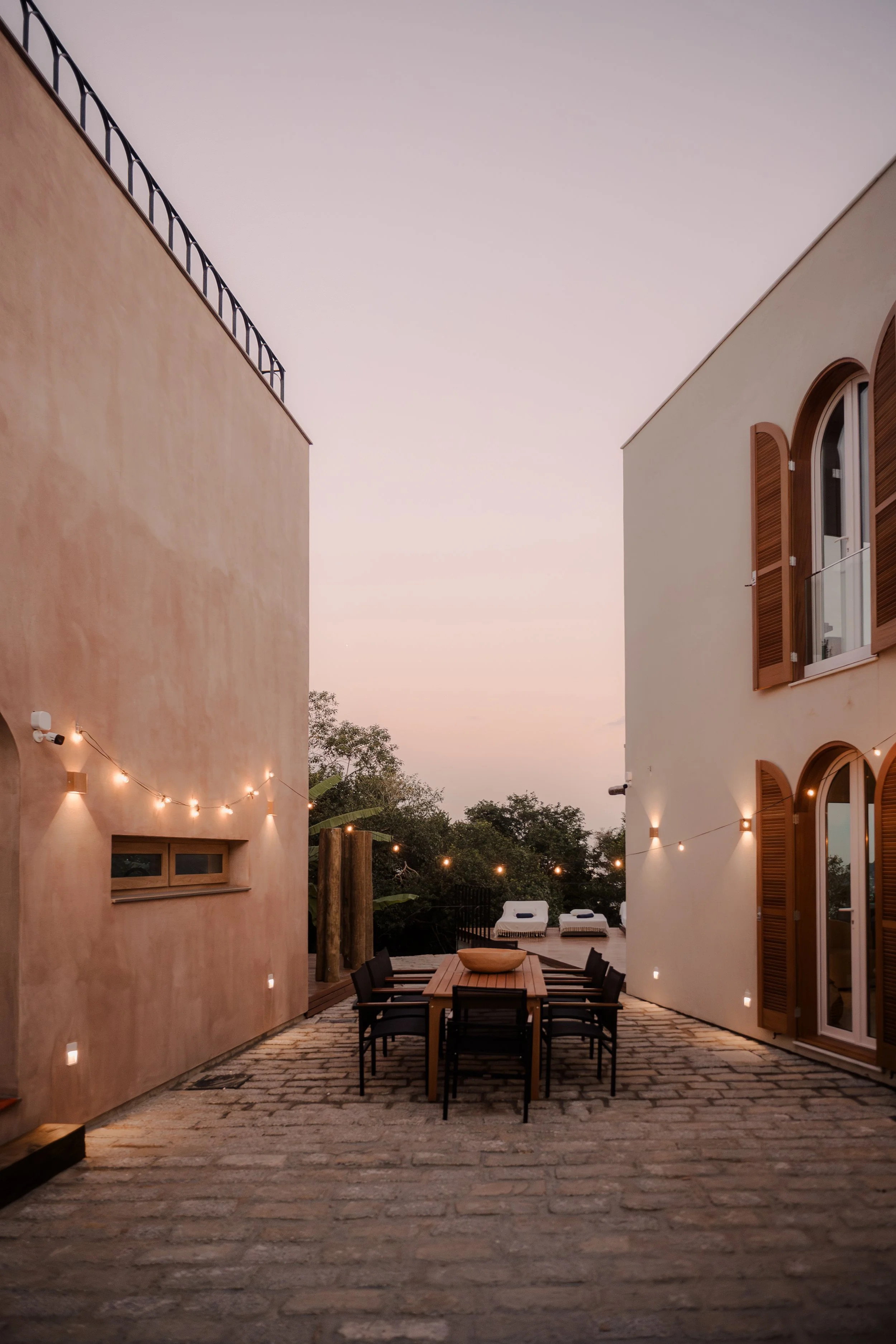 Outdoor dining area with a wooden table and black chairs, string lights hanging between two buildings at dusk, with lounge chairs and trees in the background.