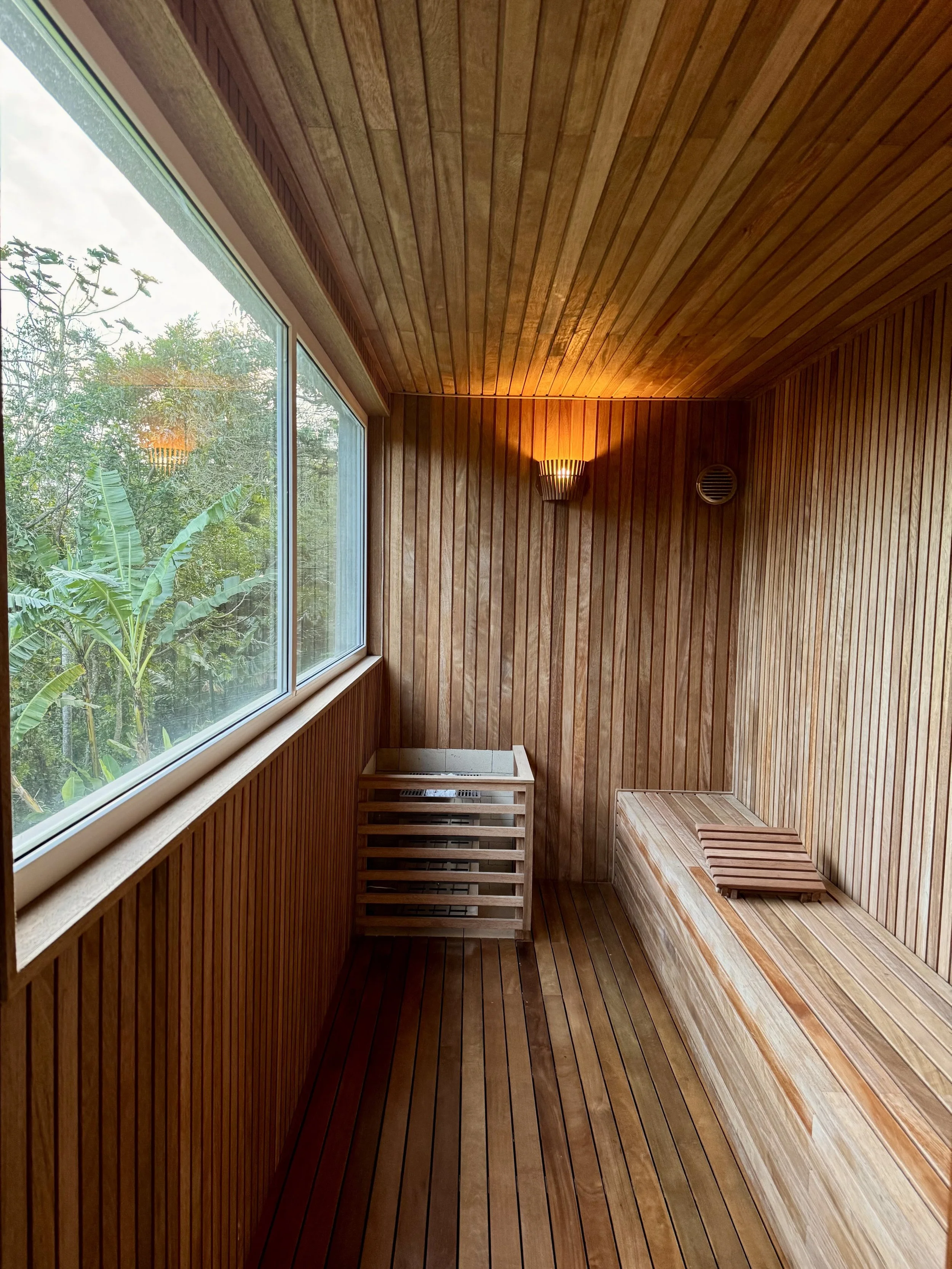 A small wooden sauna room with a large window looking out to greenery, a wooden bench, a wooden footrest, and a heater on the floor near the window, with warm lighting on the wall.