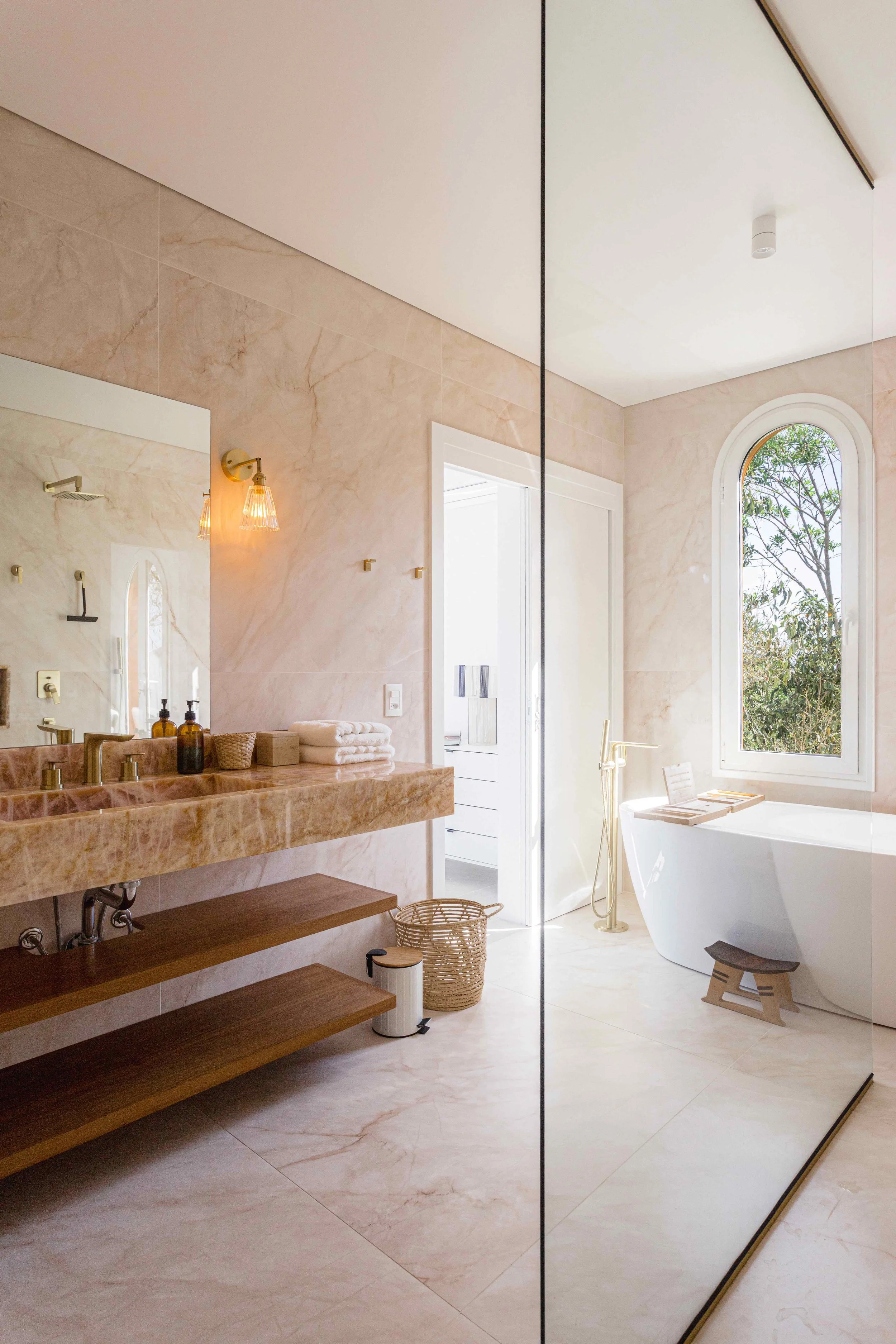 A modern bathroom with a beige marble vanity, wooden shelves, a large mirror, a wicker basket, a white bathtub by a window, and a gold floor-standing showerhead.