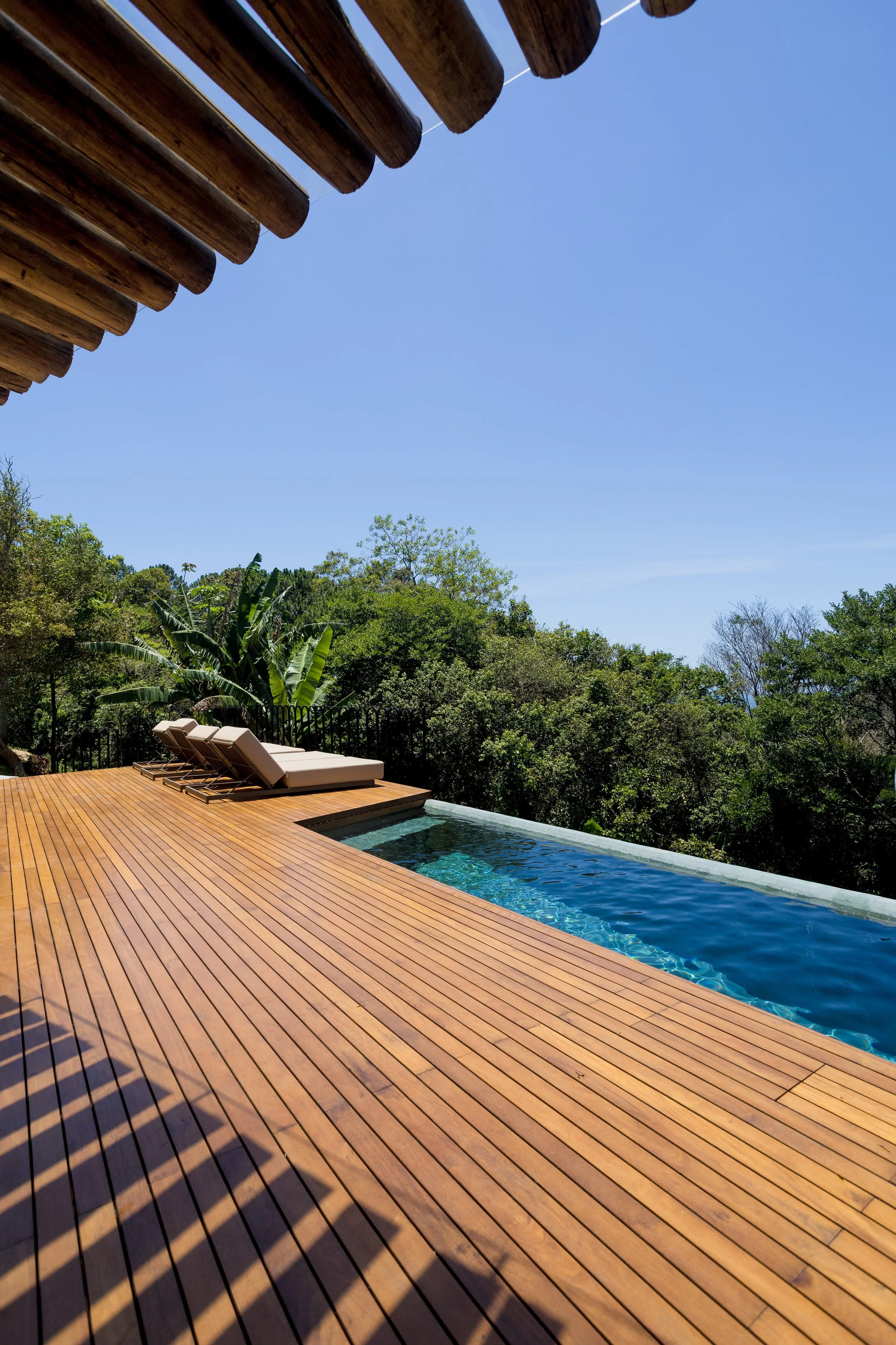 A wooden deck with lounge chairs next to an infinity pool, overlooking a lush green forest under a clear blue sky.