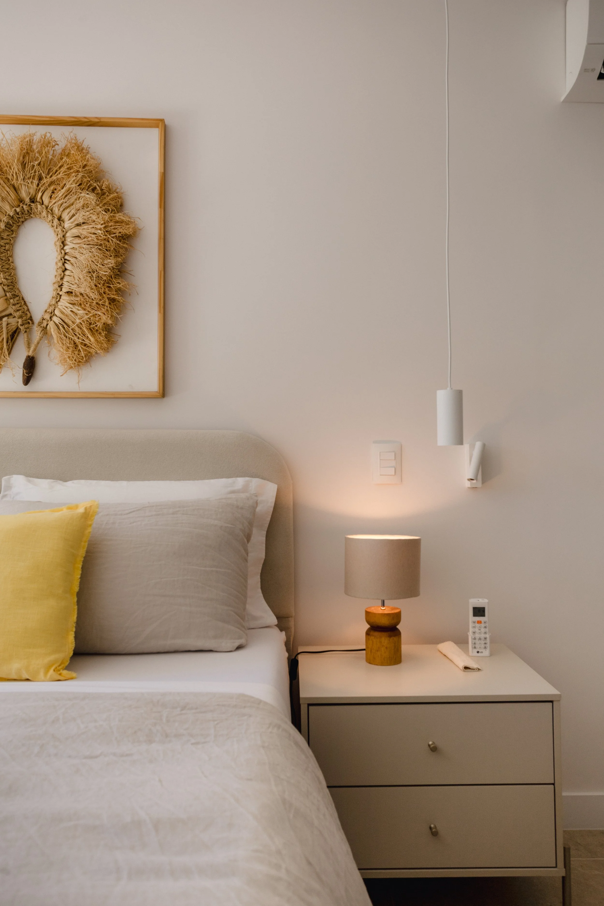 A cozy bedroom corner featuring a bed with beige and yellow pillows, a nightstand with a lamp, remote control, and rolled-up cloth, and wall decorations including a woven art piece and hanging white pendant lights.