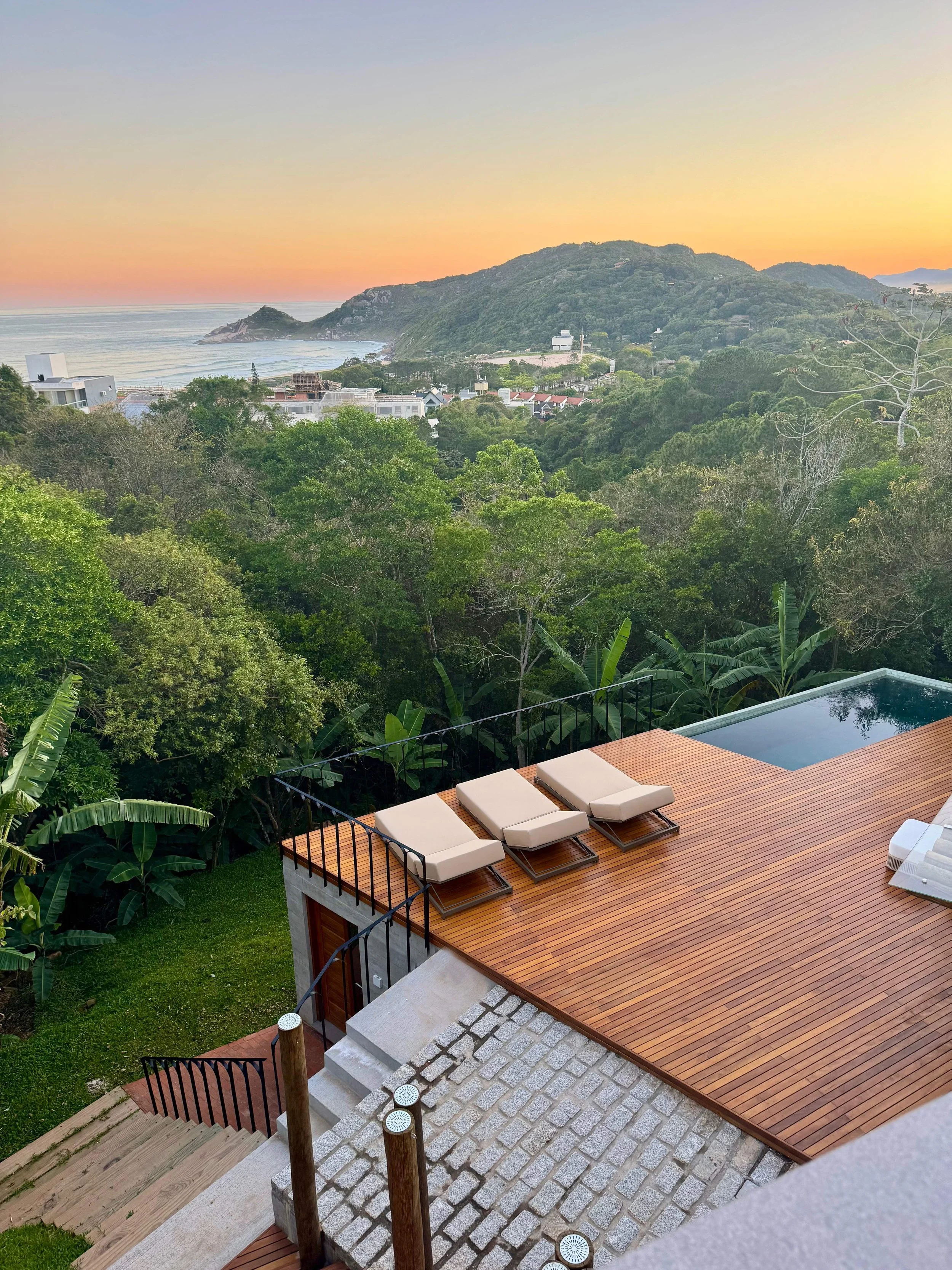 A deck with three lounge chairs and a small pool, overlooking a lush green forest, hills, and the ocean at sunset.